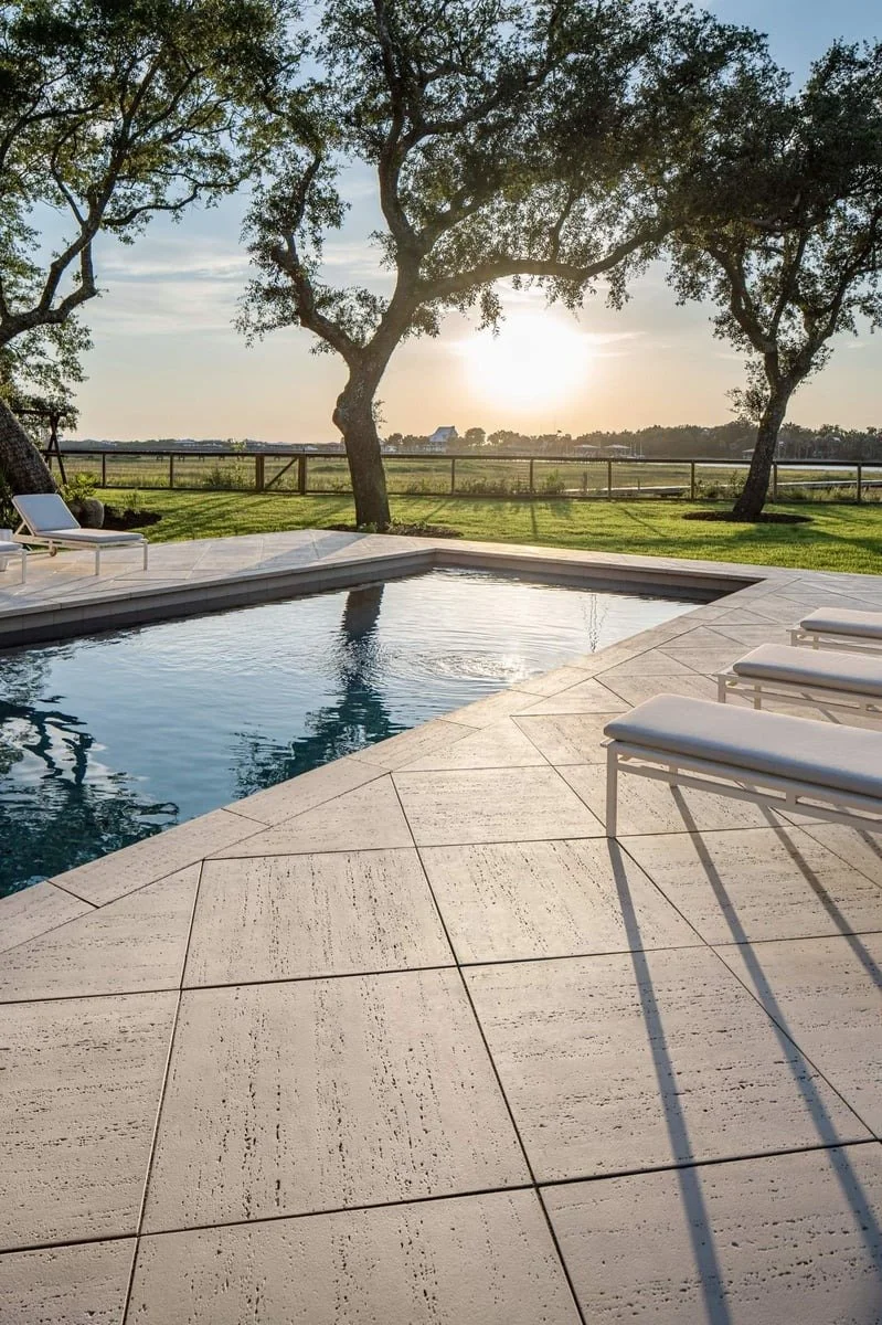 A backyard pool area at sunset with lounge chairs, trees, and a fence in the background.