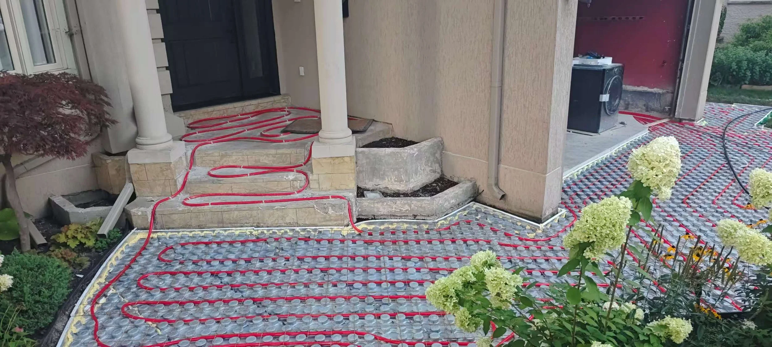 Exterior house entrance with underfloor heating system installation, visible red tubing laid out on a mesh for heating, stone steps leading up to a black door, surrounded by plants and flowers.