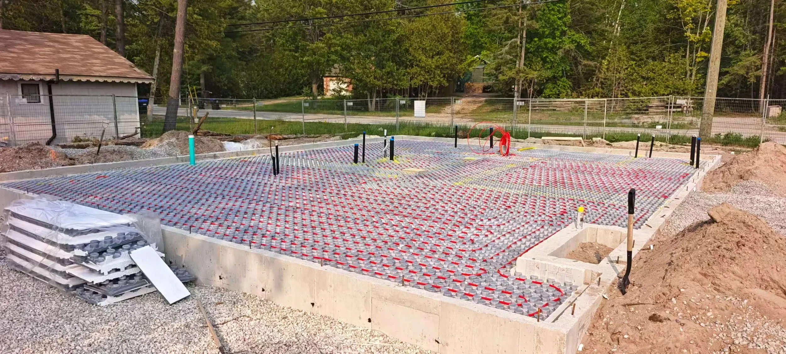 Construction site with a concrete foundation, pipe installations, and underfloor heating system with red pipes and metal plates, surrounded by dirt and construction materials.