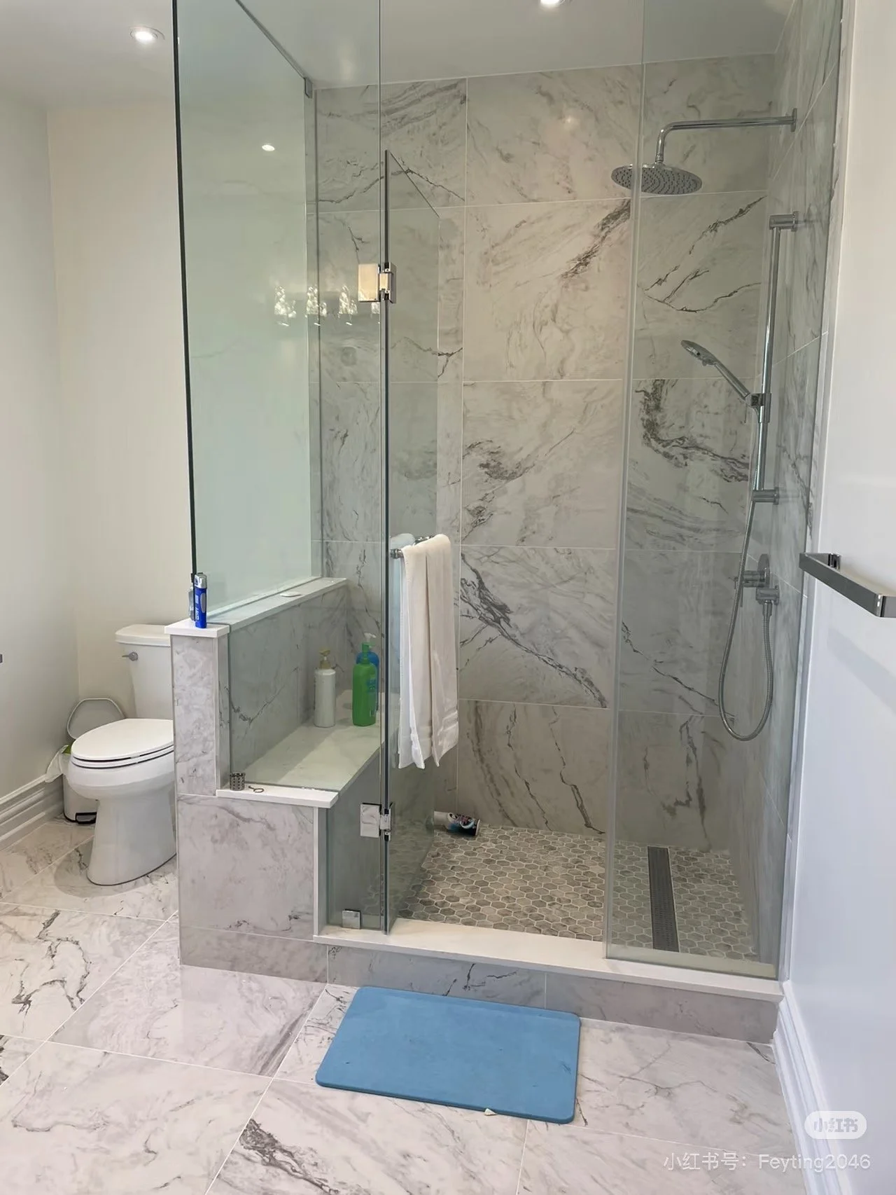 A modern bathroom with a glass-enclosed shower featuring marble tiles, a grey hexagonal tile floor in the shower, a rain showerhead, handheld shower, white toilet, and a blue bath mat outside the shower.