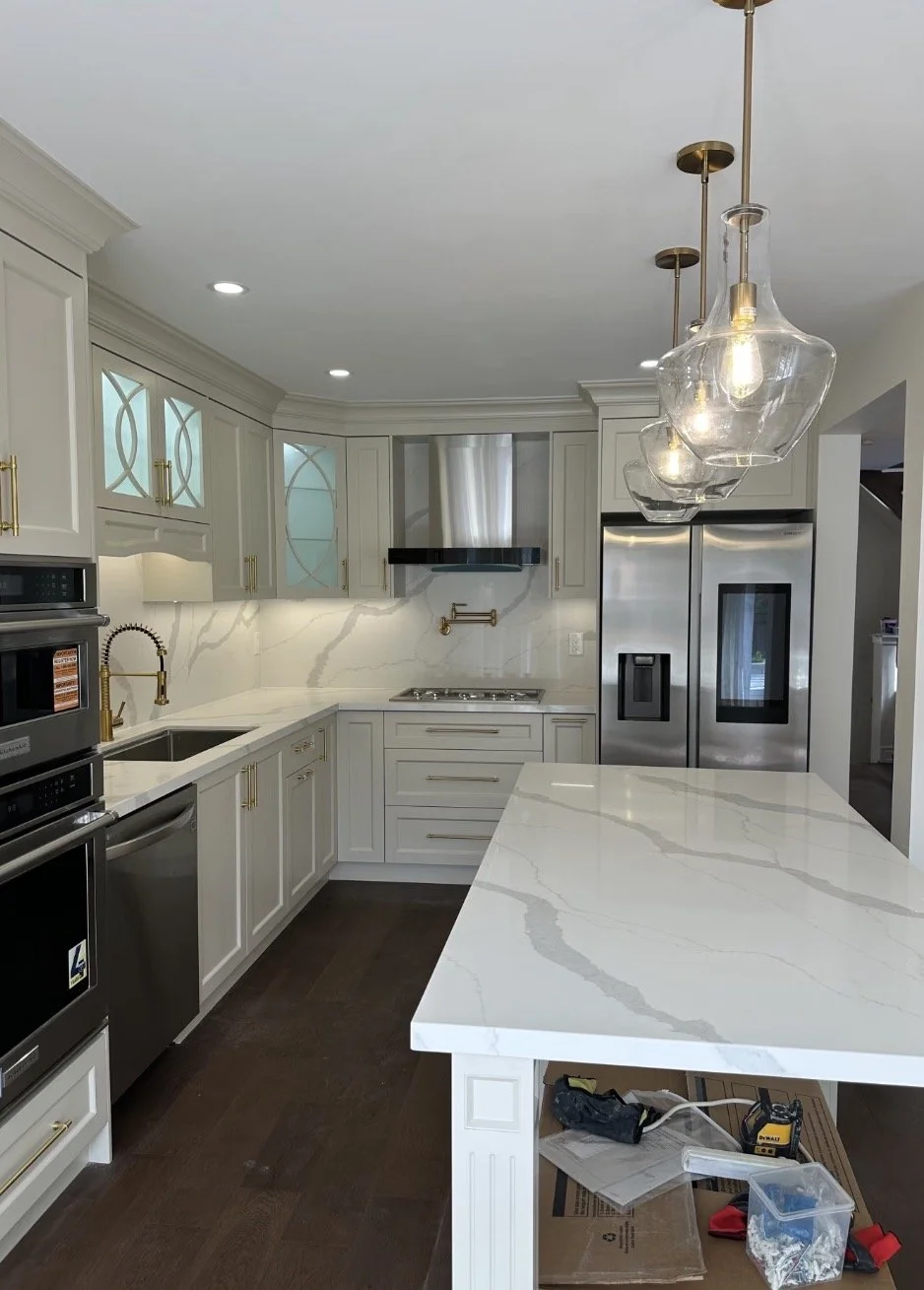 Modern kitchen with white cabinetry, gold hardware, marble countertops, stainless steel appliances, pendant lights, and hardwood floors.