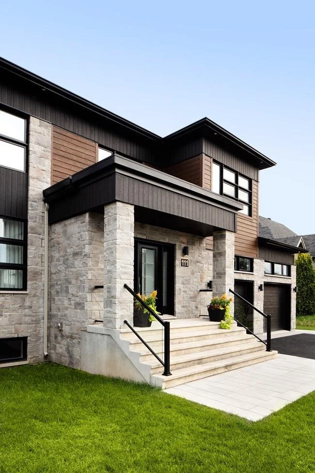 Modern two-story house with stone and wood exterior, front porch with stairs, black railings, and a garage on the right, surrounded by a green lawn under a clear sky.