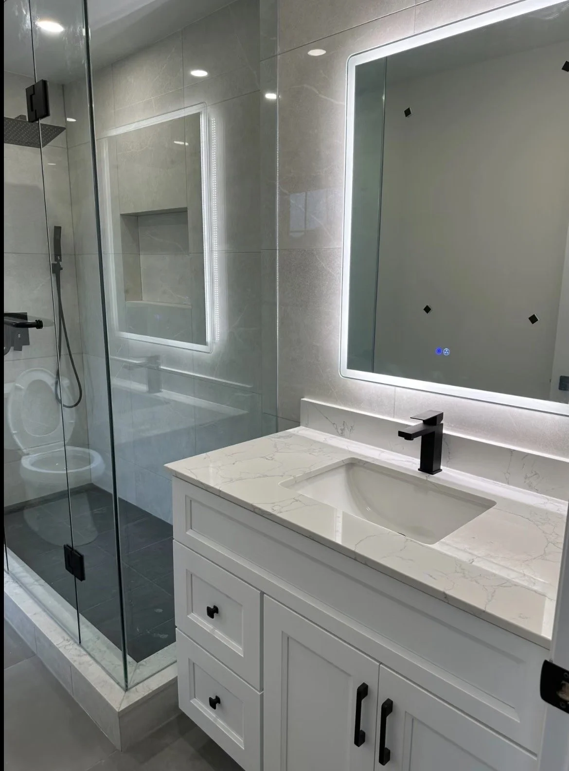 Modern bathroom with a glass shower enclosure, a white vanity with a marble countertop, and a mirrored backlit medicine cabinet.