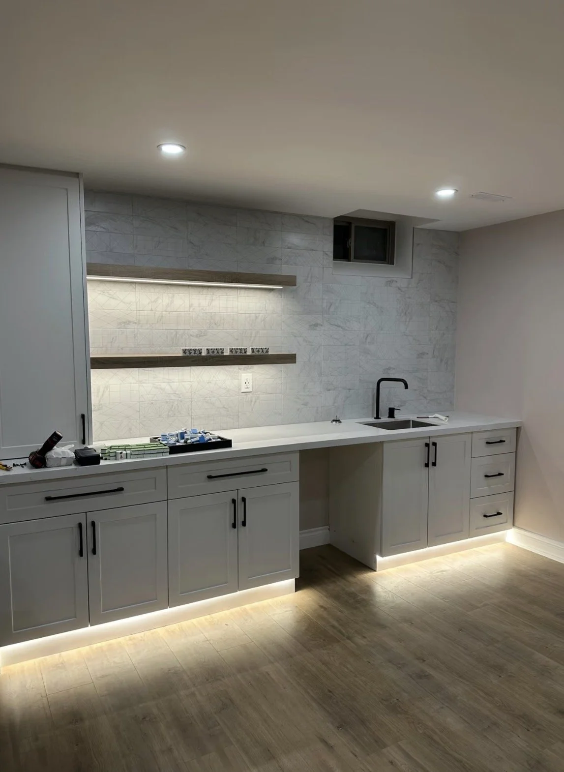 A modern kitchen with white cabinetry, black hardware, a black faucet, gray backsplash tiles, and wooden floors. Under-cabinet lighting illuminates the space.