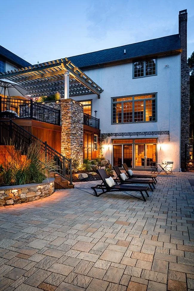 Exterior view of a modern two-story house with outdoor seating area, stone patio, and a pergola at dusk.