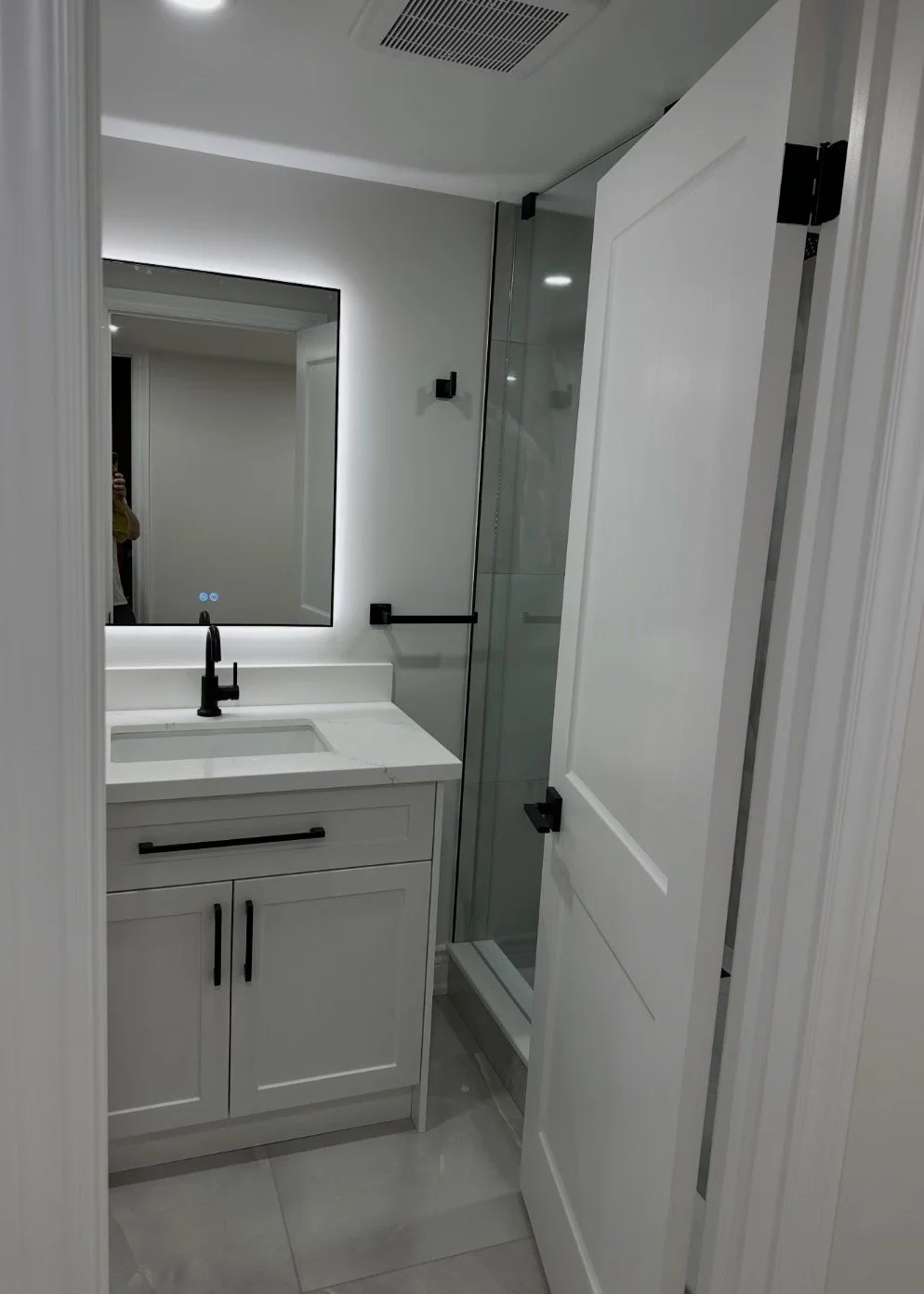 A modern bathroom featuring a white vanity with black handles, a black faucet, a mirror with backlit lighting, a glass-enclosed shower, and white walls and floor tiles.