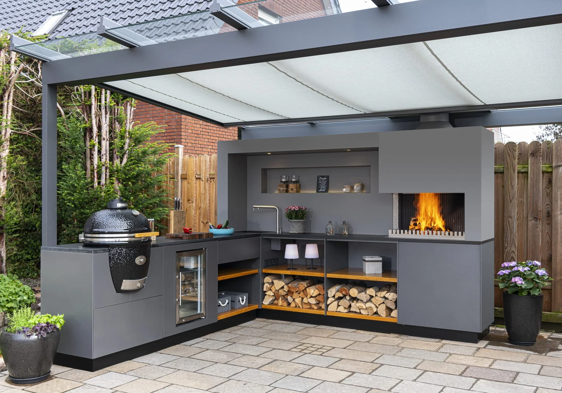 Outdoor kitchen with a built-in fireplace, wood storage, and a barbecue grill on a patio with potted plants and a covered roof.
