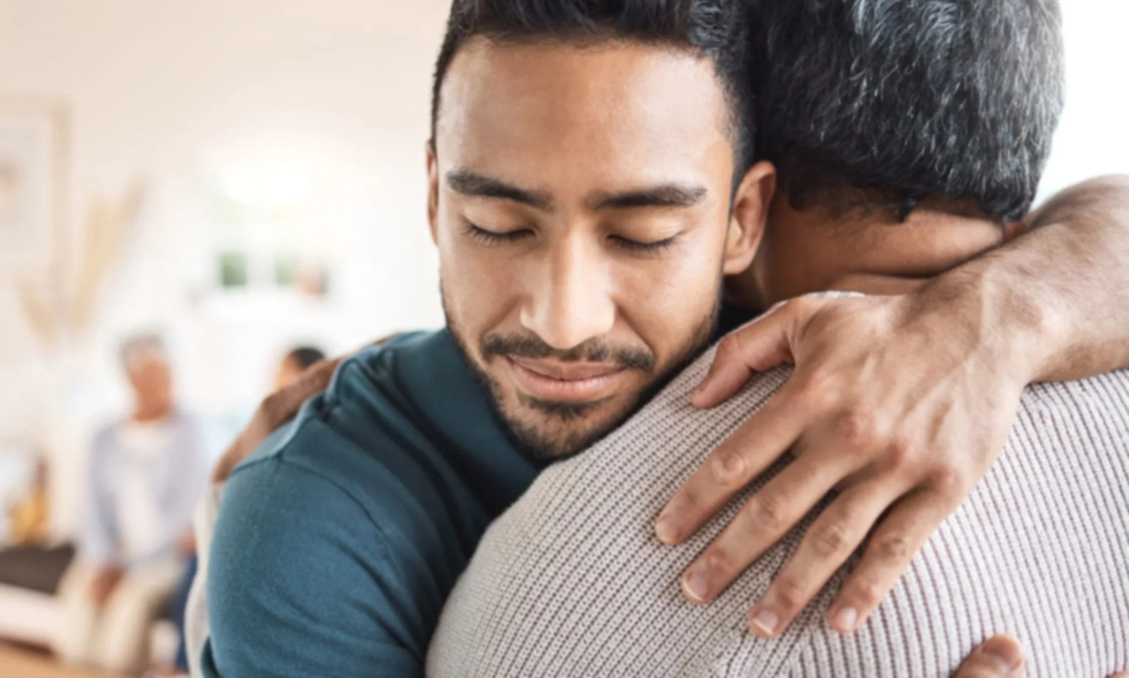 An image of an older black man being embraced by a younger black man who's holding him and closing his eyes.