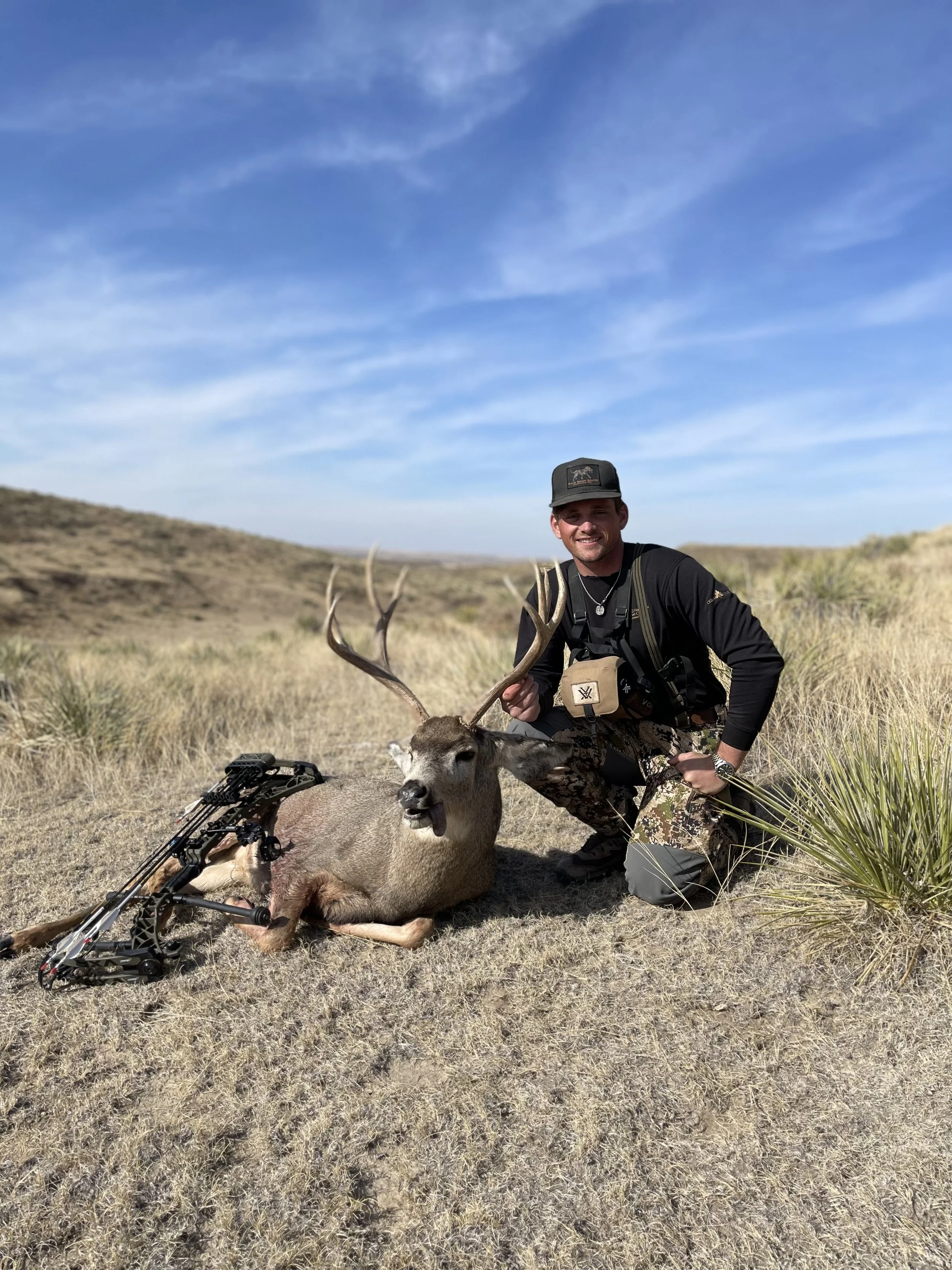 A man kneeling next to a dead deer with large antlers, holding its antlers, in a grassy, open landscape under a blue sky. A bow is lying on the ground beside the deer.
