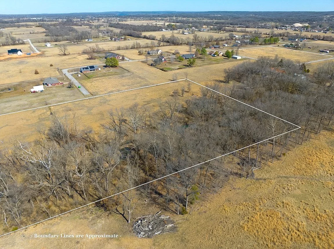 Aerial view of a rural area with a rectangular wooded lot outlined by boundary lines, surrounded by open fields and scattered houses.