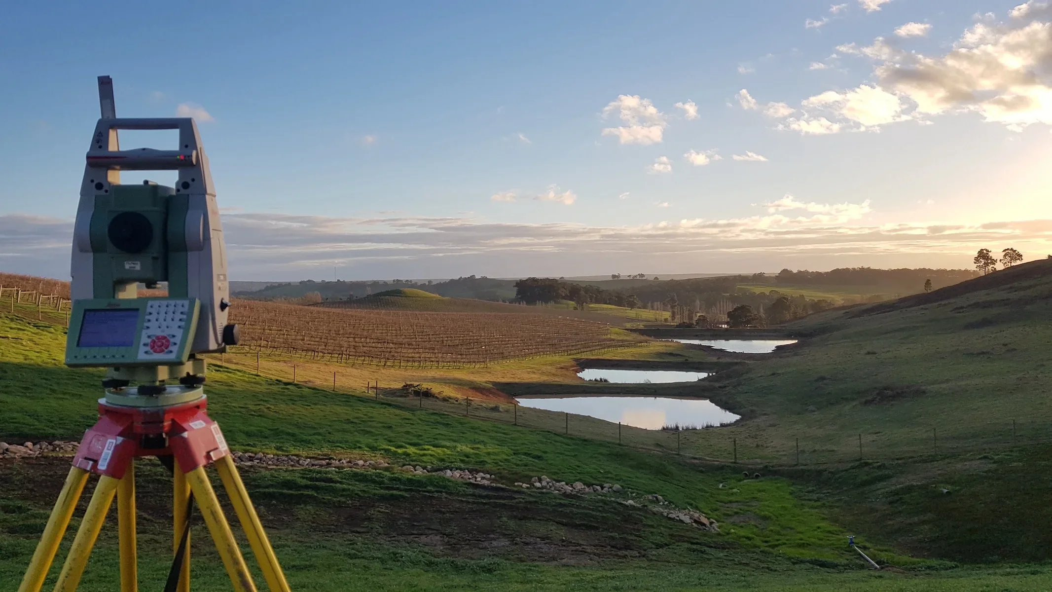 Surveying instrument set up in a rural landscape with rolling hills, ponds, trees, and a cloudy sky at sunset.