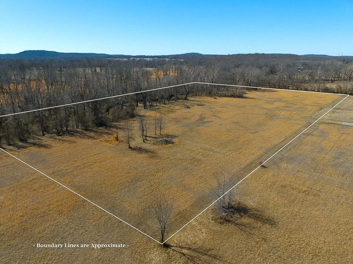 Aerial view of a large open field with sparse trees, surrounded by a wooded area, with blue sky overhead.