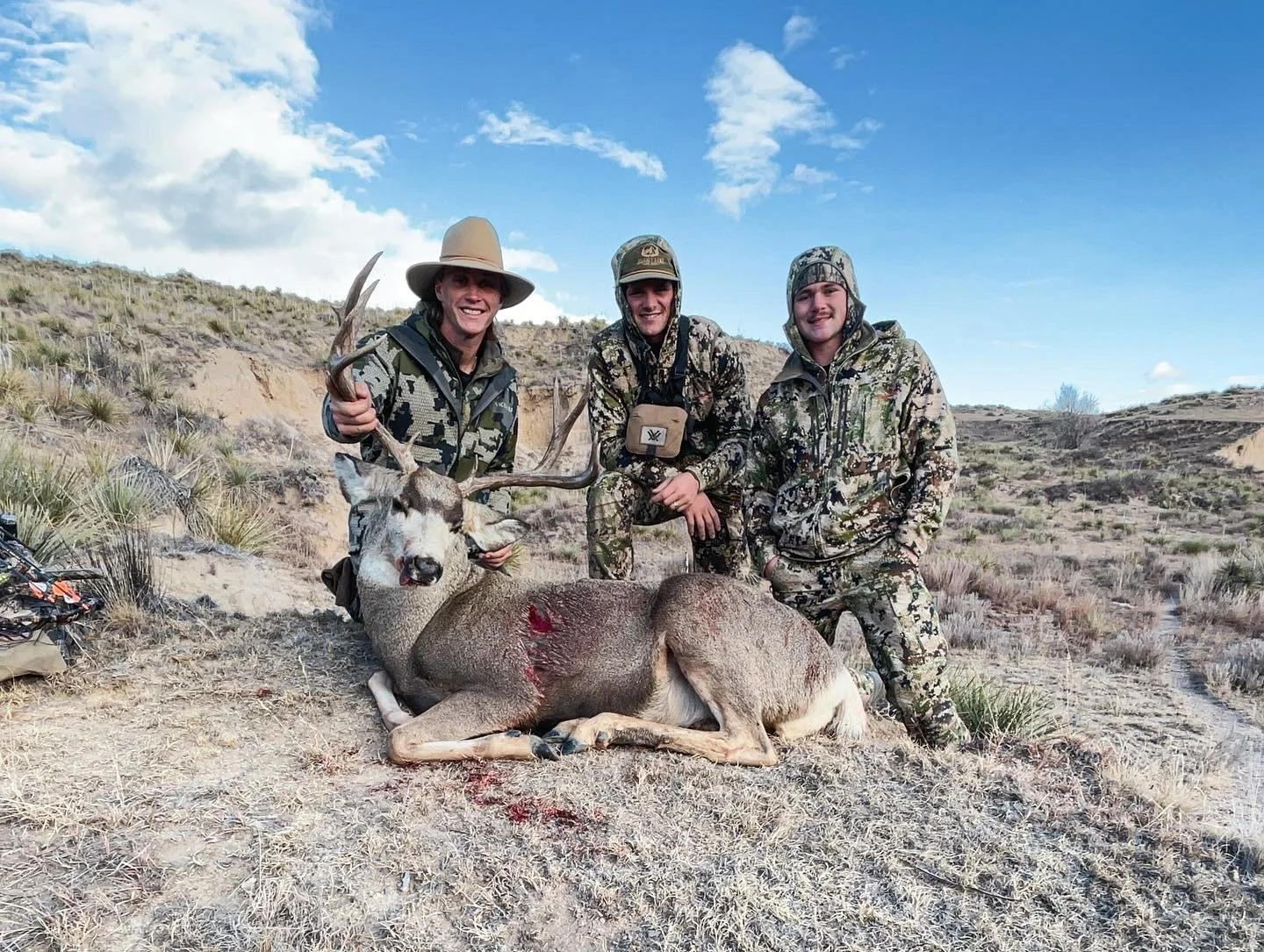 Three hunters in camouflage gear posing with a large dead deer with antlers in a dry, hilly landscape under a blue sky with scattered clouds.