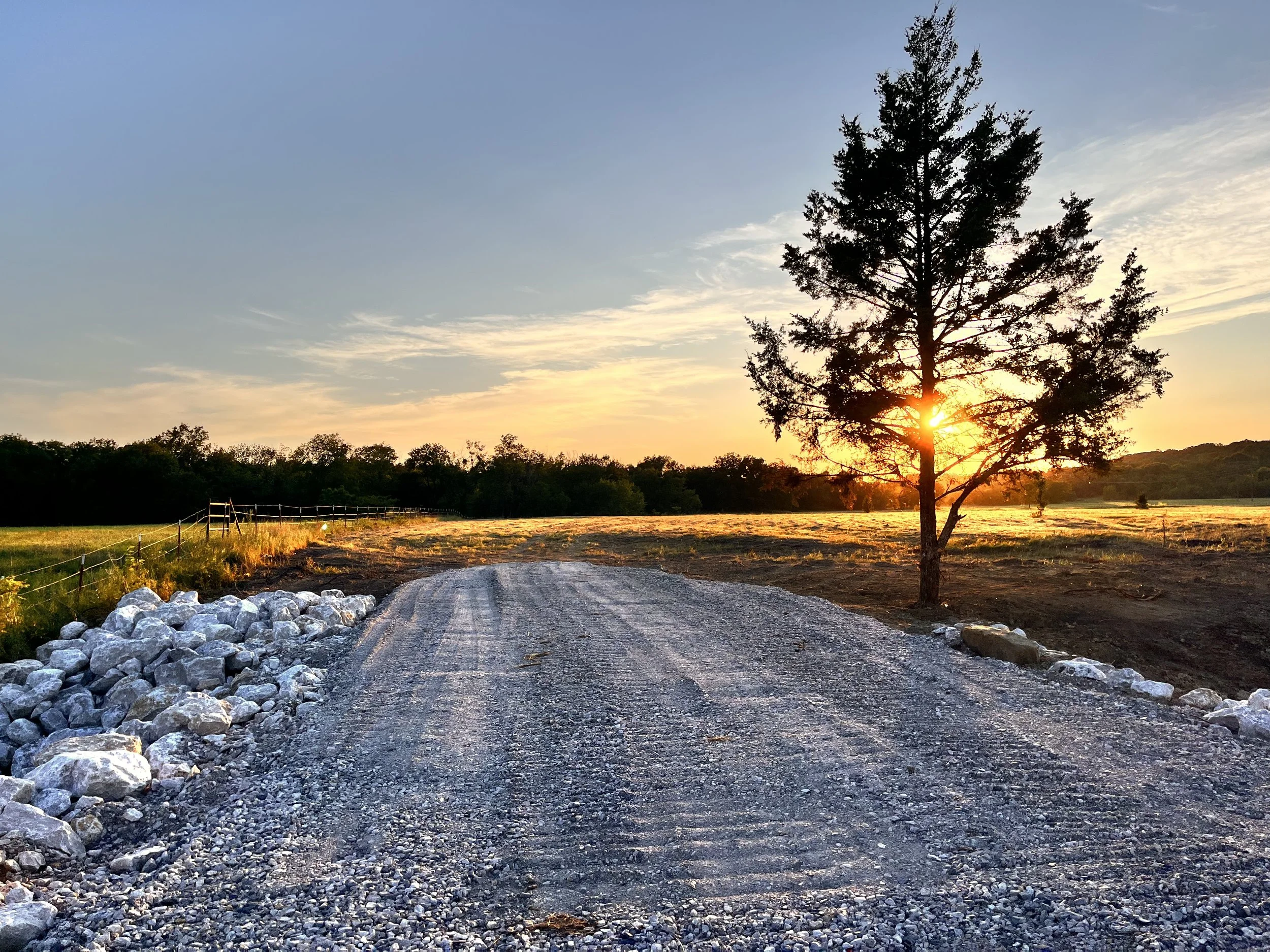 Sunset over a gravel road with a tree on the right and a fence on the left, in a rural landscape.