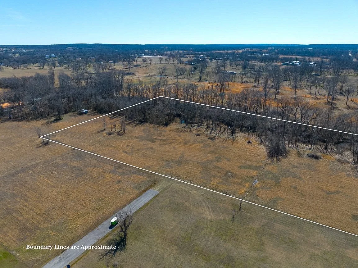 Aerial view of a large, rural, fenced-in property with open fields, trees, and a few small buildings in the distance under a clear blue sky.