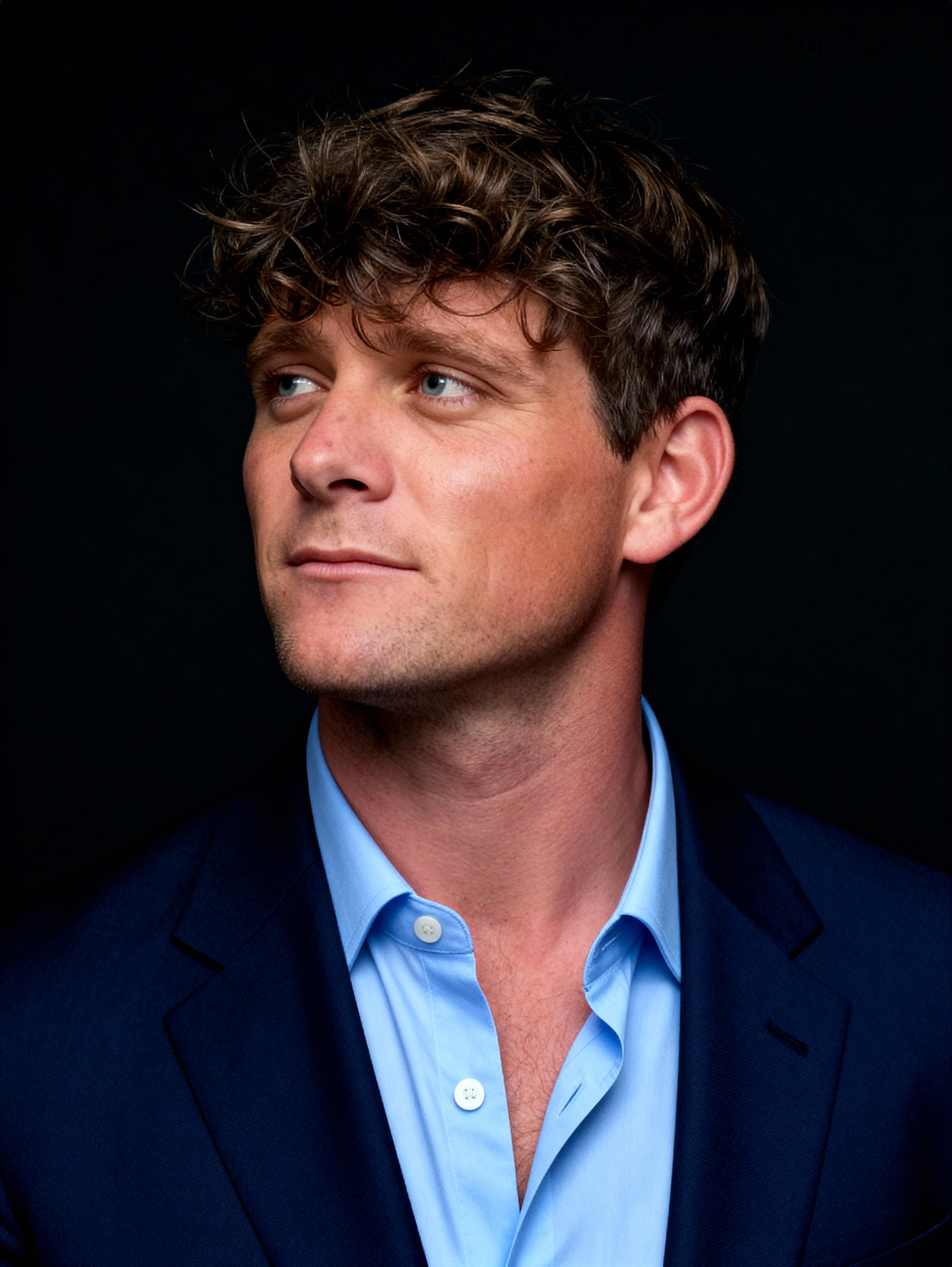Headshot of a man with curly brown hair, blue eyes, wearing a blue shirt and dark blazer, looking to the side against a dark background.