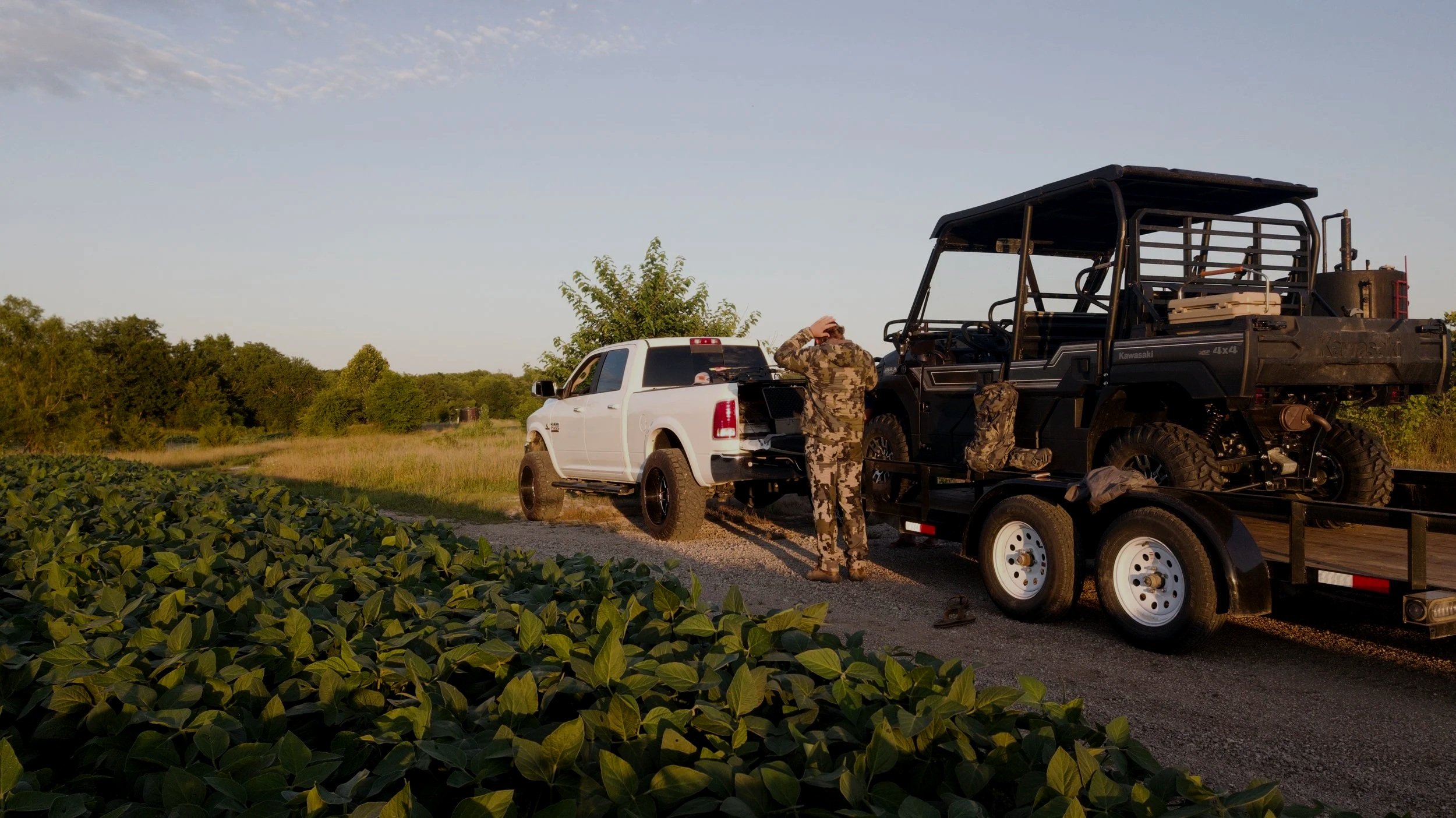 A person in camouflage clothing standing next to a white pickup truck and a trailer carrying a black off-road vehicle, set on a rural dirt road with lush green vegetation and trees in the background.
