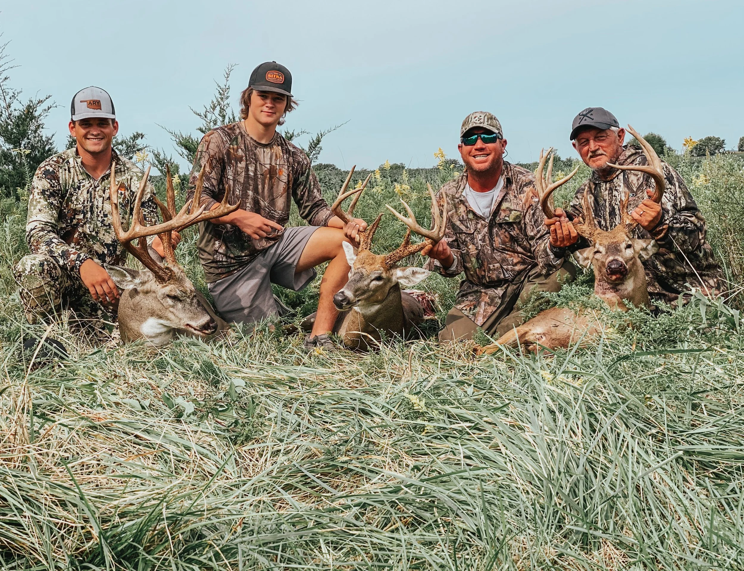 Four men posing with three deer they hunted, holding the antlers of the deer in an outdoor field with tall grass and bushes.
