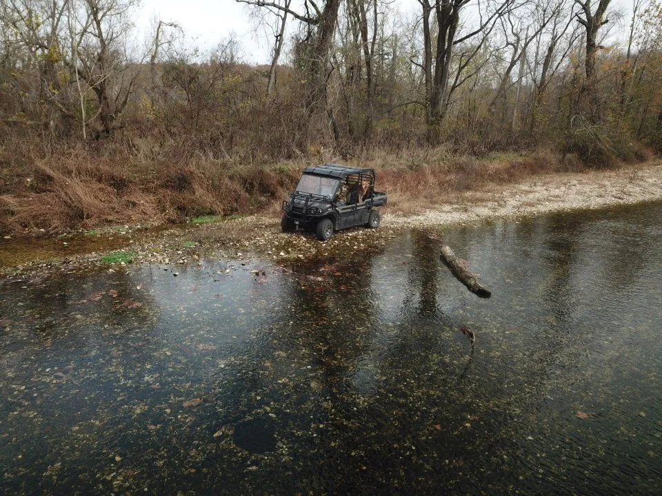 A black utility vehicle parked on the rocky edge of a shallow river, surrounded by leafless trees during fall.