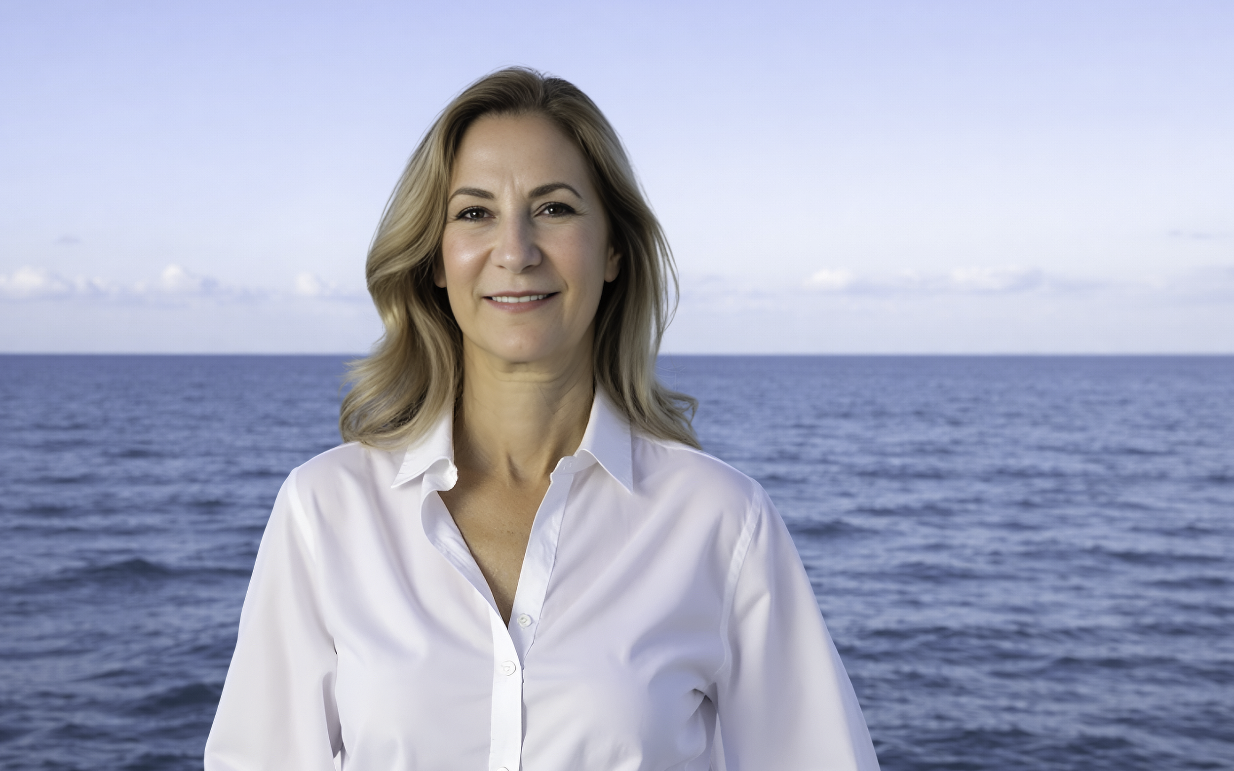 A woman with blonde hair wearing a white shirt standing by the ocean with blue sky and clouds in the background.