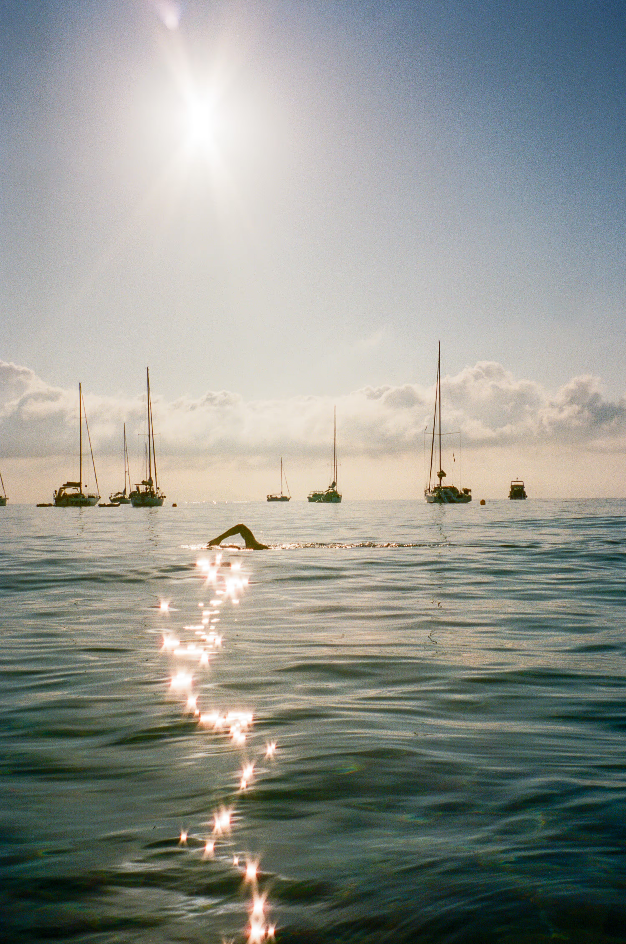A person swimming in the ocean near sailboats under the bright sun.