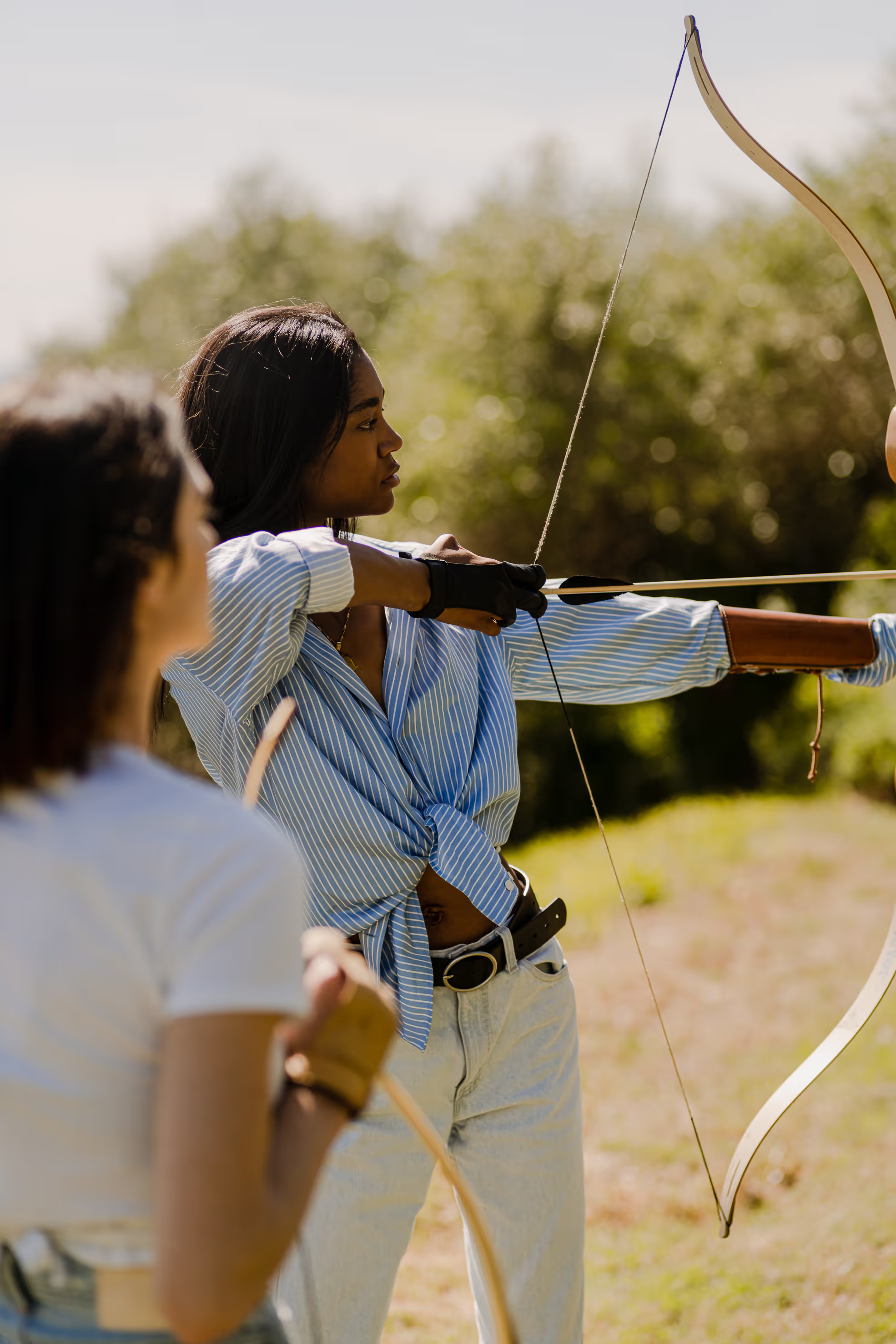 A woman wearing a blue striped shirt is practicing archery outdoors, drawing a bow and aiming, with another person in the foreground.