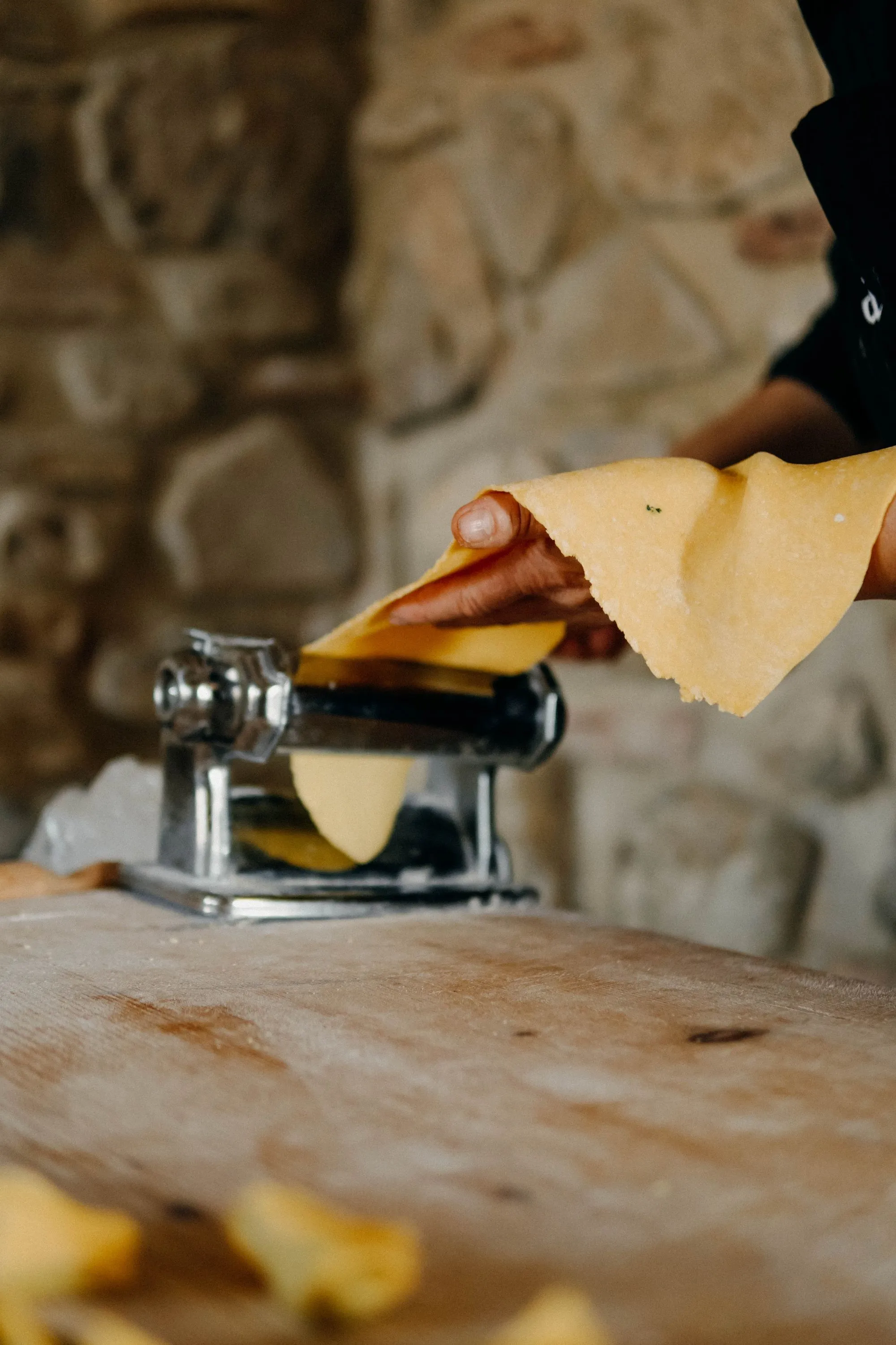 A person uses a pasta machine to roll out fresh pasta dough on a wooden surface, with a stone wall in the background.