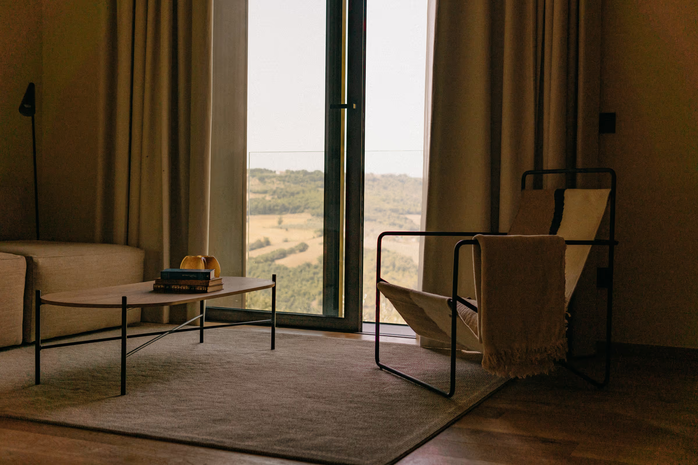 Living room with open glass door showing scenic countryside view, beige couch, modern coffee table with books and a decorative object, armchair covered with a blanket, and curtains.