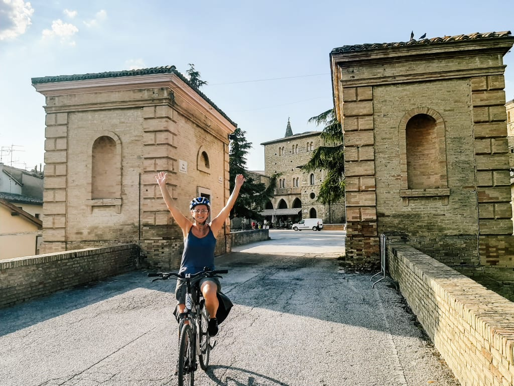 Woman on bicycle with arms raised, historic stone buildings in the background, on a sunny day.