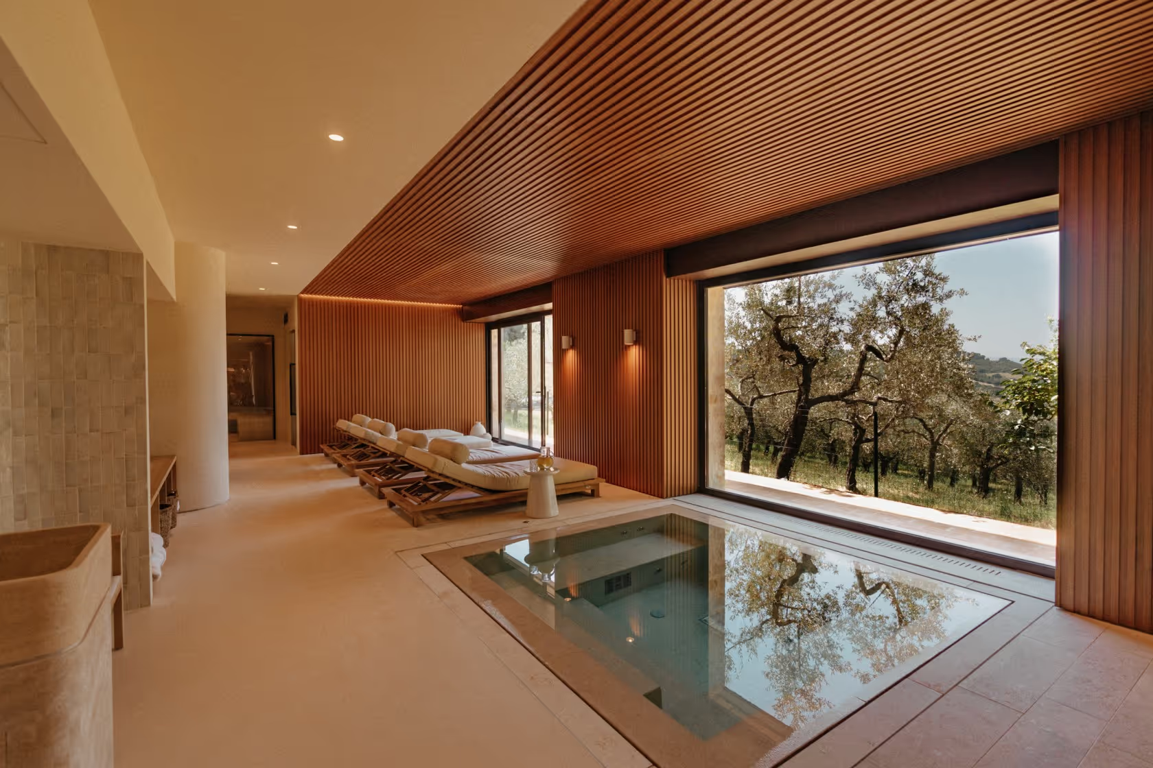 Luxury indoor spa room with a heated plunge pool, wooden lounge chairs with cushions facing large glass windows overlooking a tree-filled landscape, and soft warm lighting.