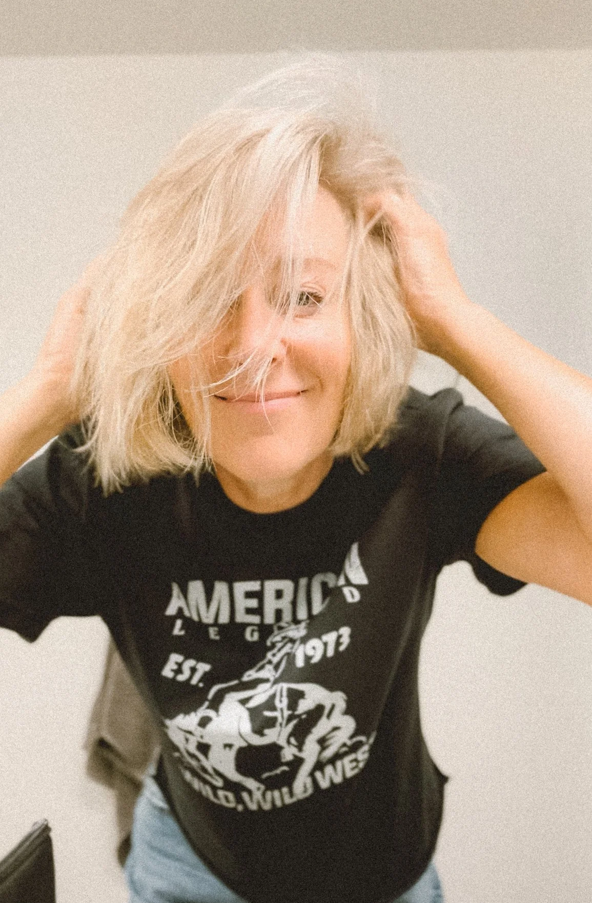 A woman with shoulder-length blonde hair, smiling, wearing a black T-shirt with white graphic text and a graphic of a horse, standing against a plain background.