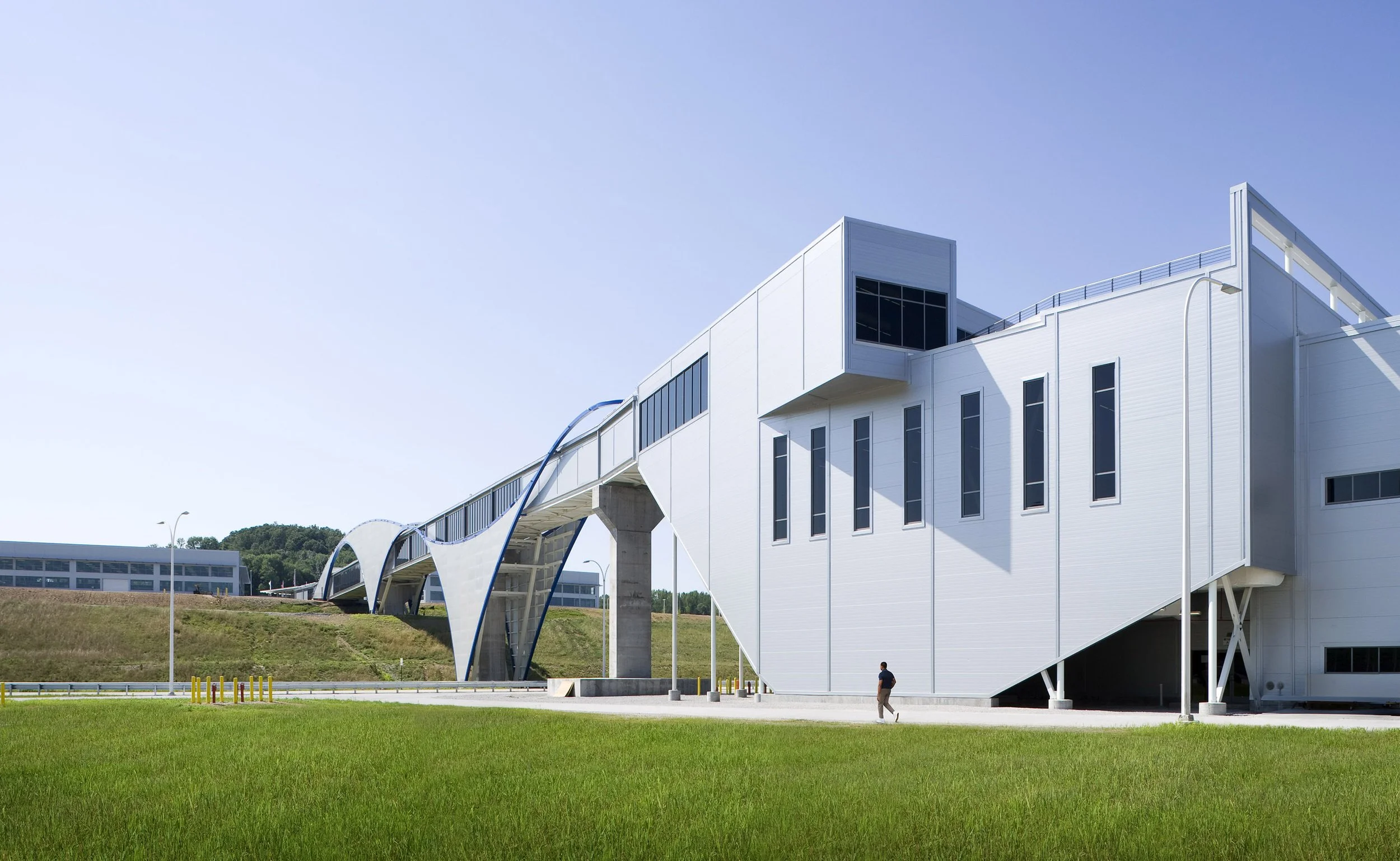 Modern white building with geometric design, connected by a curved walkway, set on a grassy area with a person walking nearby under a clear blue sky.
