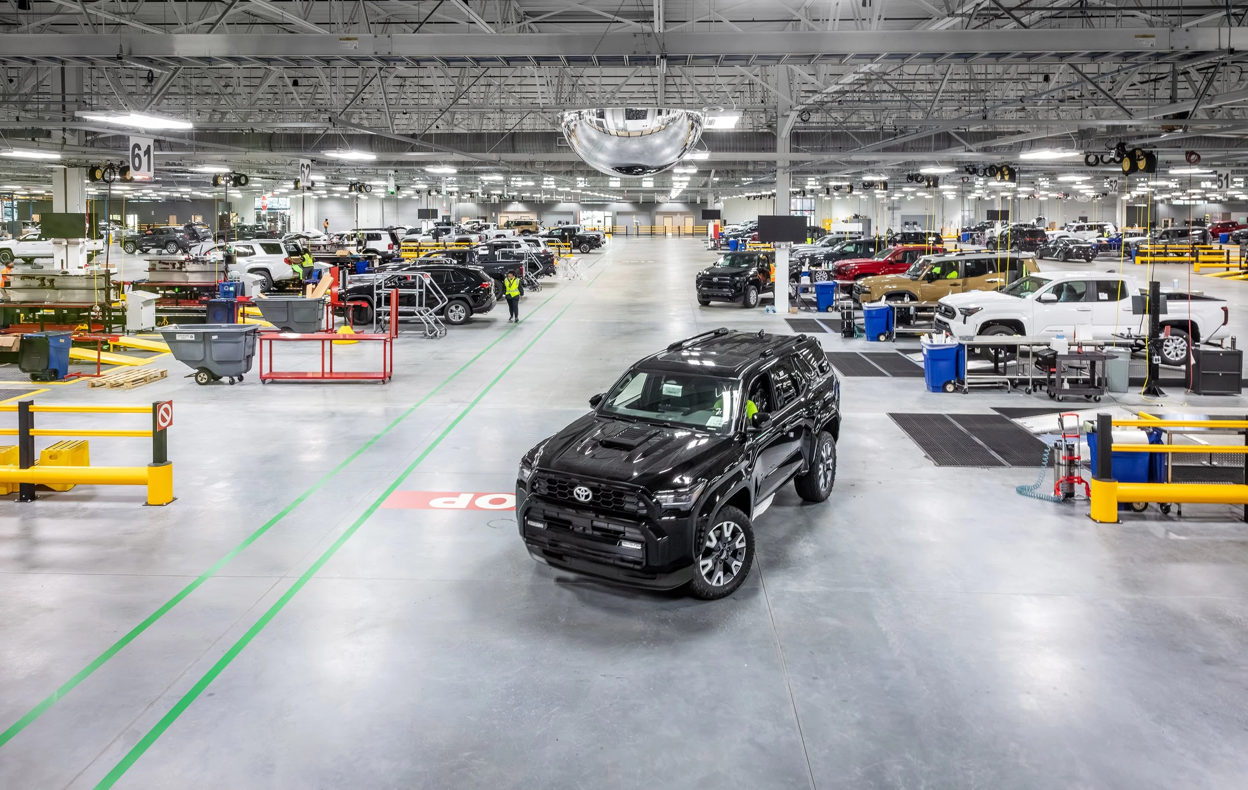 A large, brightly lit automotive factory with a black SUV in the center, surrounded by multiple vehicles being assembled and workers in safety vests working throughout the spacious facility.