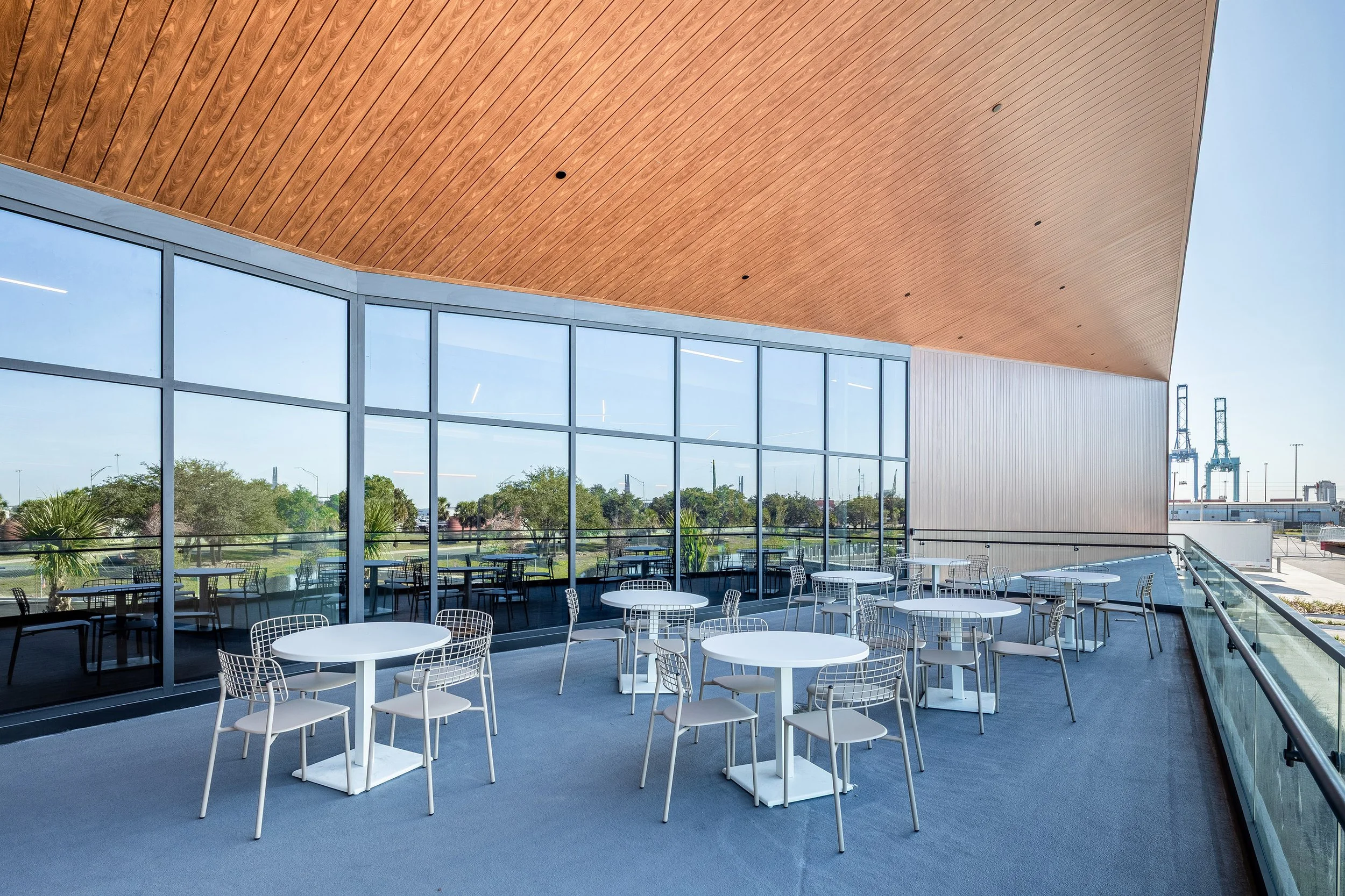 A modern outdoor terrace with round white tables and wire chairs, set on a blue floor. Large glass windows reflect the sky and trees, and a wooden ceiling extends overhead. The space overlooks a distant industrial area.