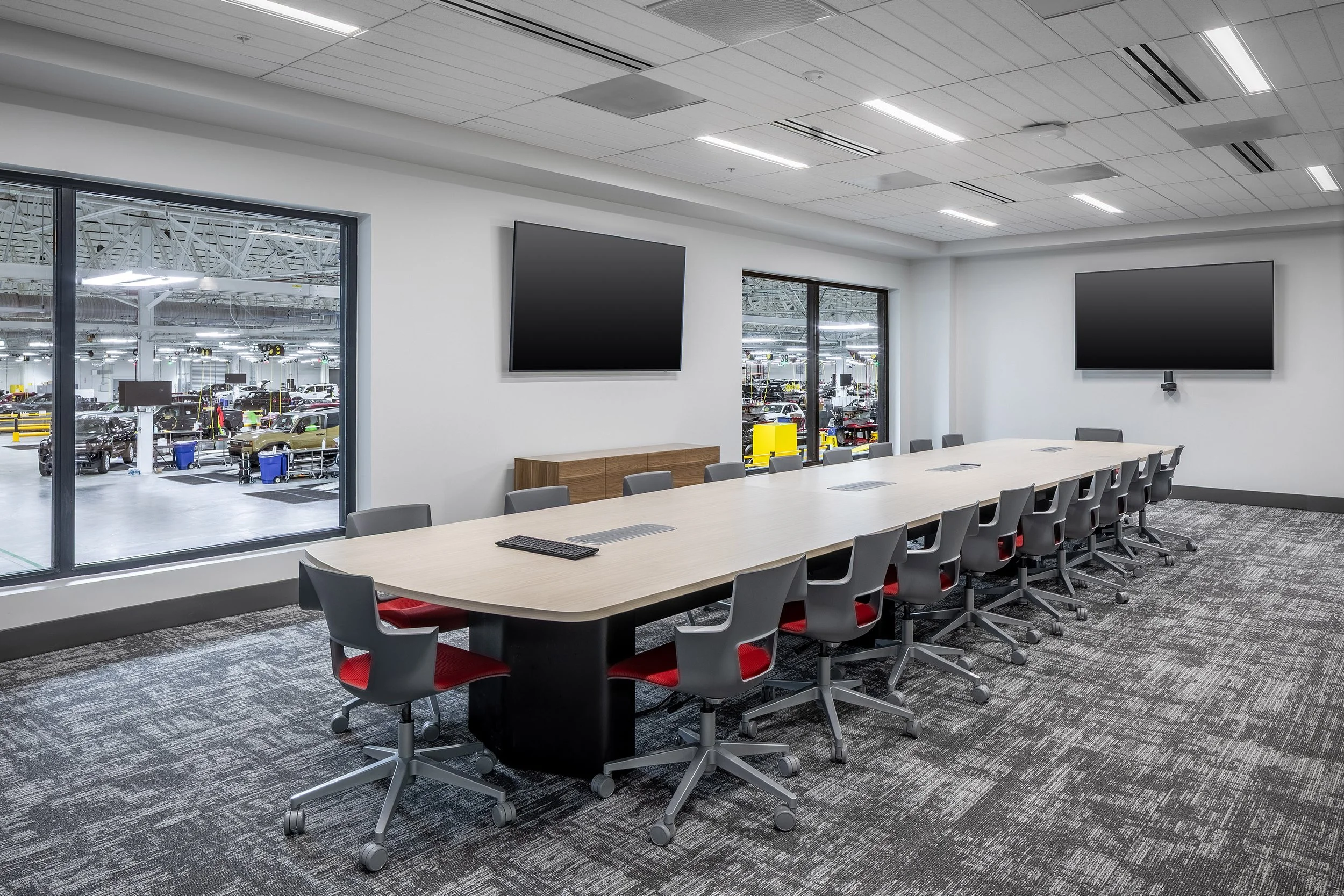 Modern conference room with a long table, gray and red chairs, two large wall-mounted screens, and windows overlooking a factory floor with industrial equipment. The room has gray patterned carpet and white walls.