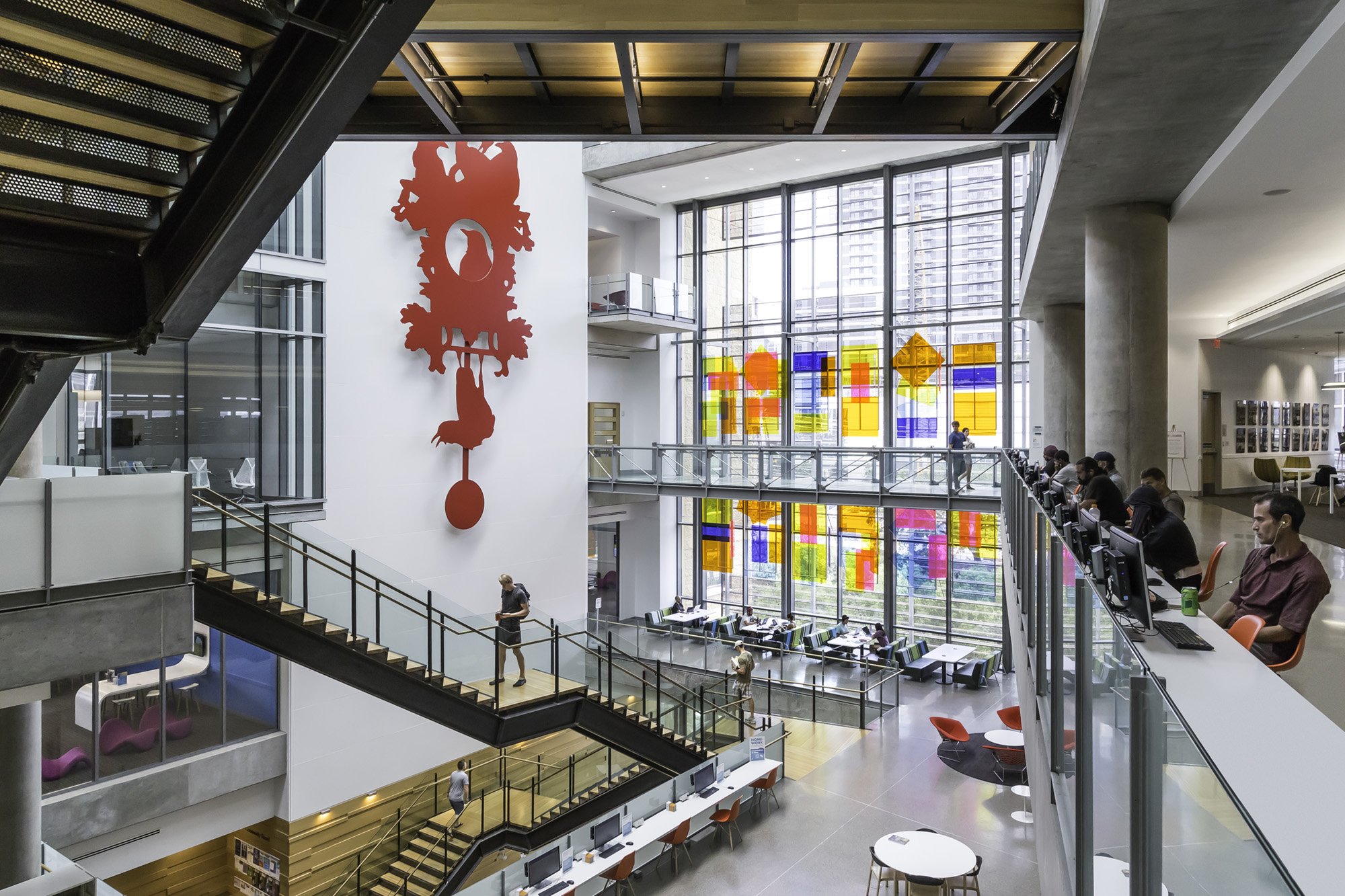 Modern multi-level interior of a Austin Public Library with large windows, colorful stained glass art, and people working and walking. A large red abstract sculpture hangs on the wall.