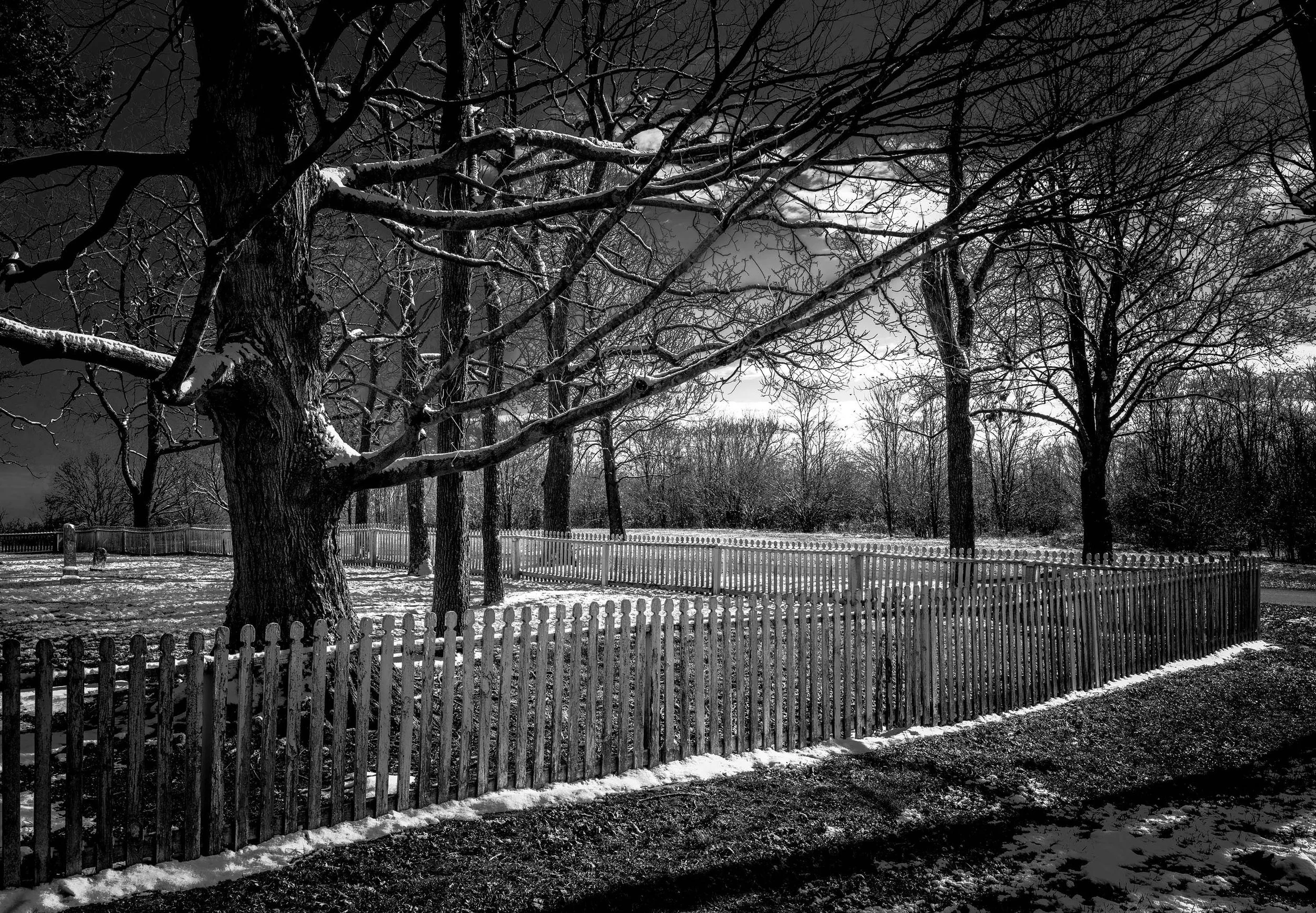 Black and white photo of bare trees, a snowy fenced landscape, and  cloudy sky in park near historic building.