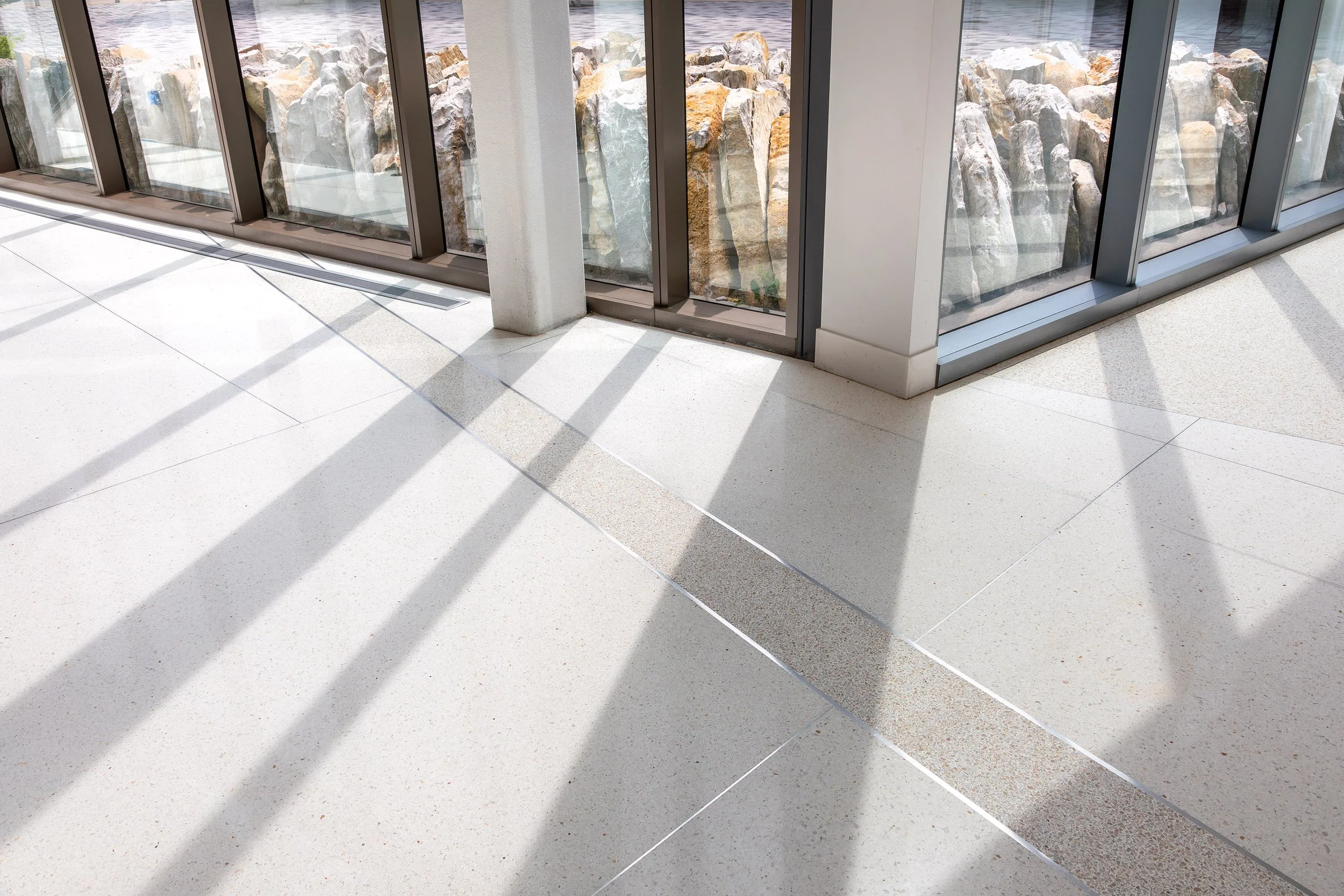 Sunlight casts window frame shadows onto a cream-colored terrazzo floor beside large glass windows overlooking rocks outside.