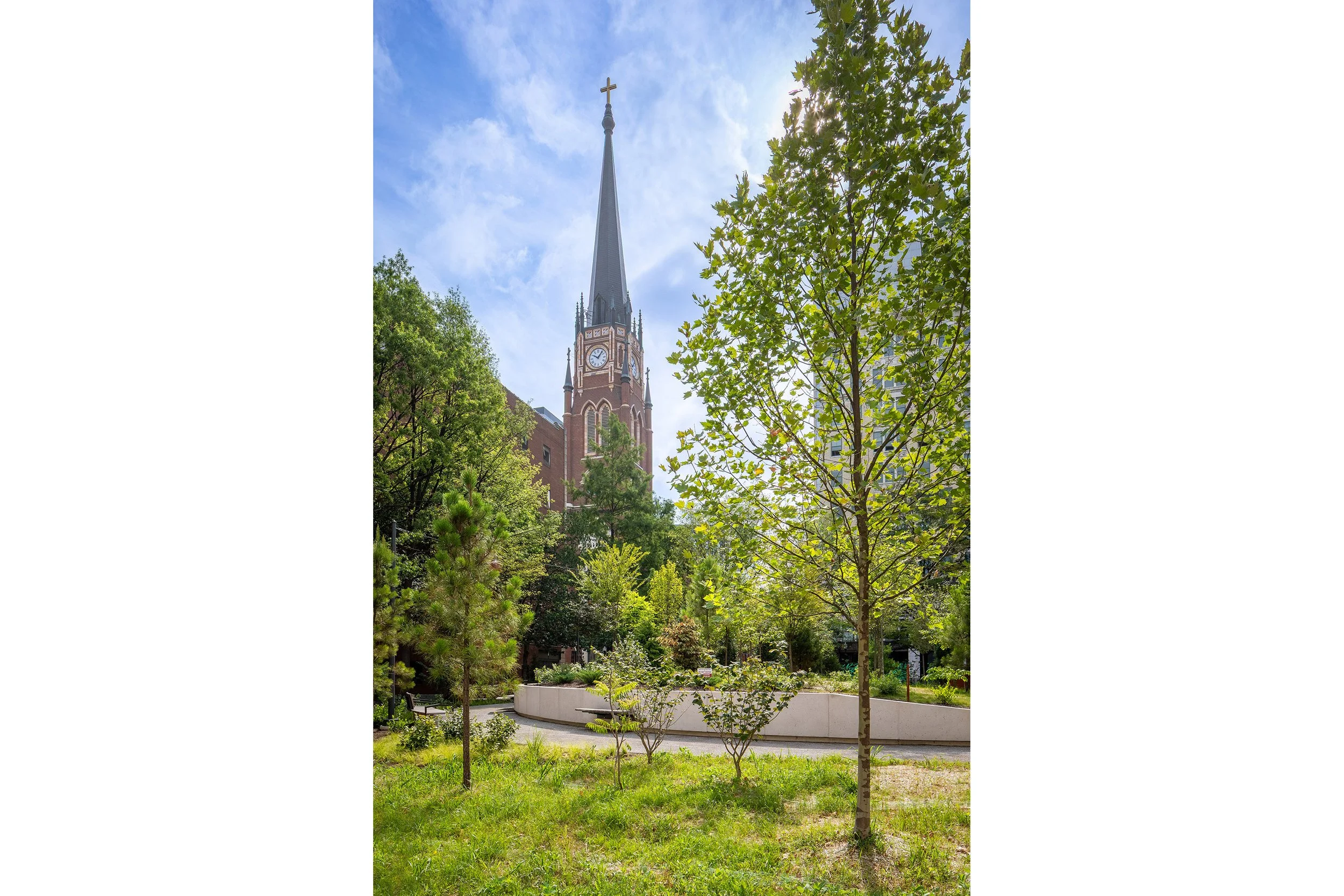 A tall, pointed church steeple with a cross rises above lush green trees and a grassy park area, creating a striking piece of architecture set against a blue sky with scattered clouds.