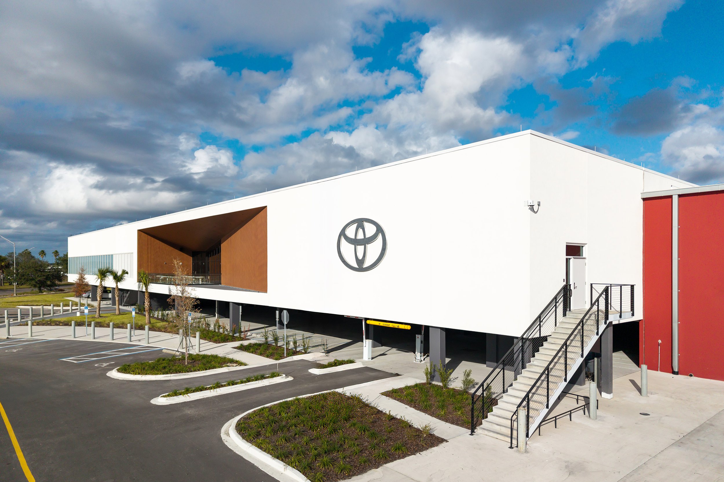 A modern white building with the Toyota logo on the facade, featuring an external staircase and a mostly empty parking lot, under a partly cloudy sky.