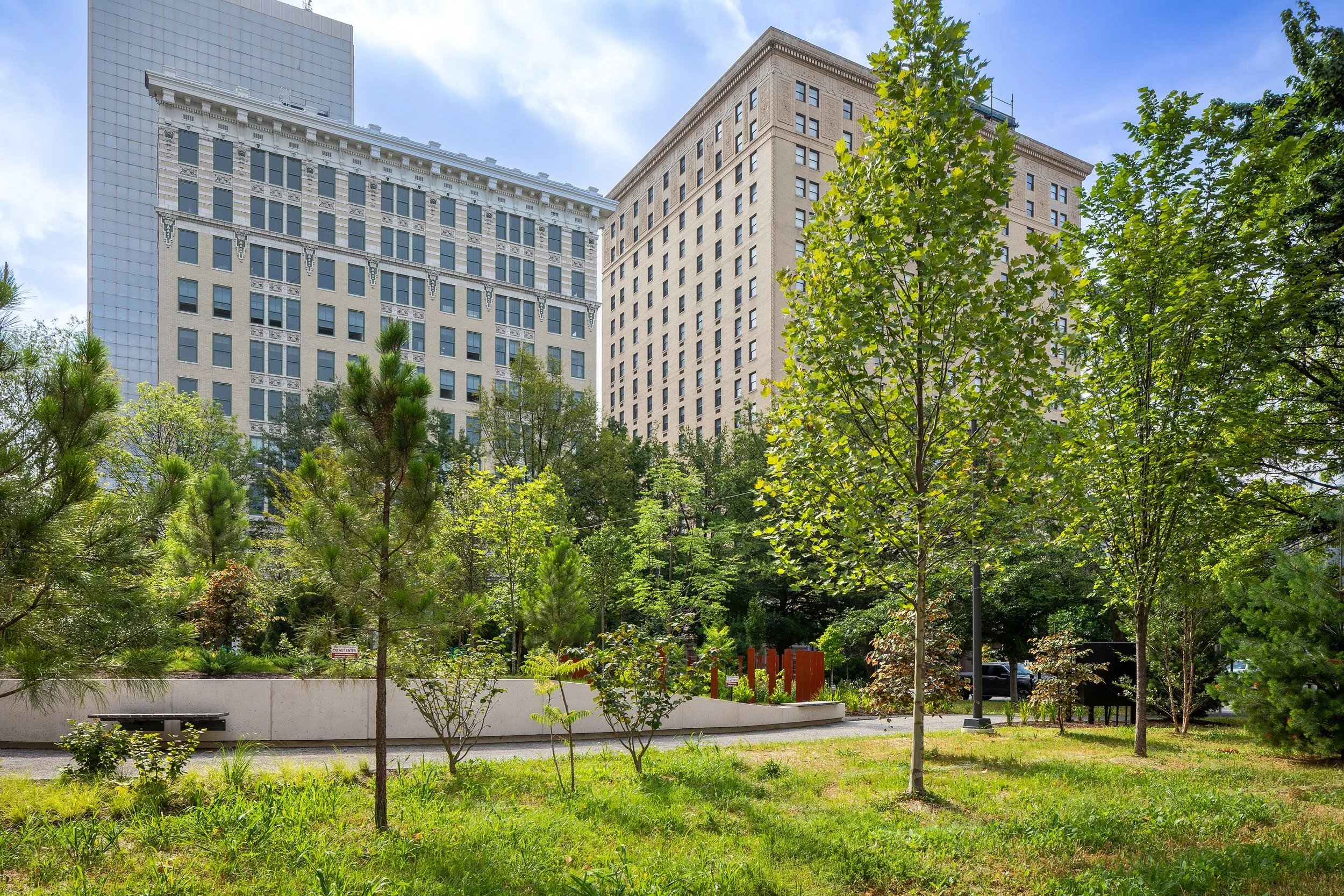A small urban park with green trees and grass, surrounded by tall office buildings under a partly cloudy sky.
