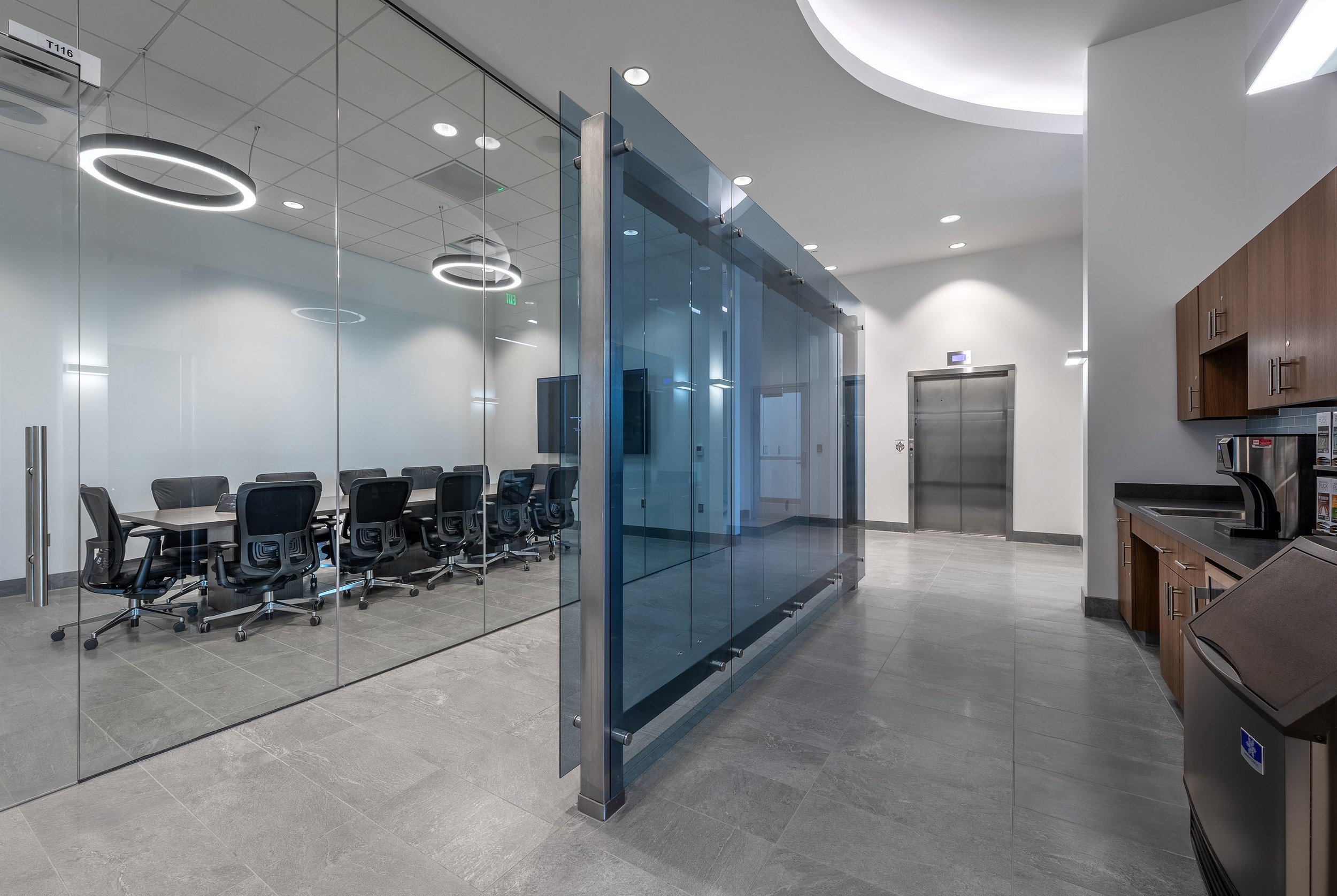 Modern office space with a glass-walled conference room on the left, black chairs around a table, elevators in the back, and a kitchenette on the right. Bright lighting and sleek gray flooring highlight its architectural style—perfect for photography