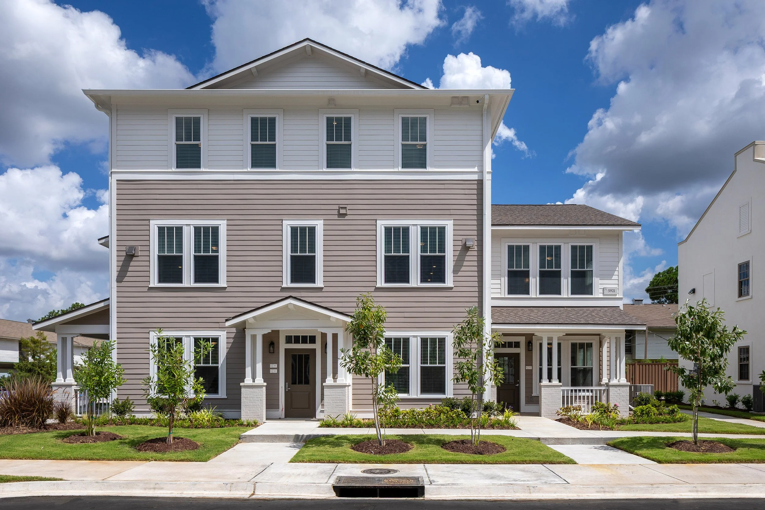 A modern three-story townhouse with gray and white exterior, large windows, and a small green lawn with young trees in front, under a bright blue sky with scattered clouds.
