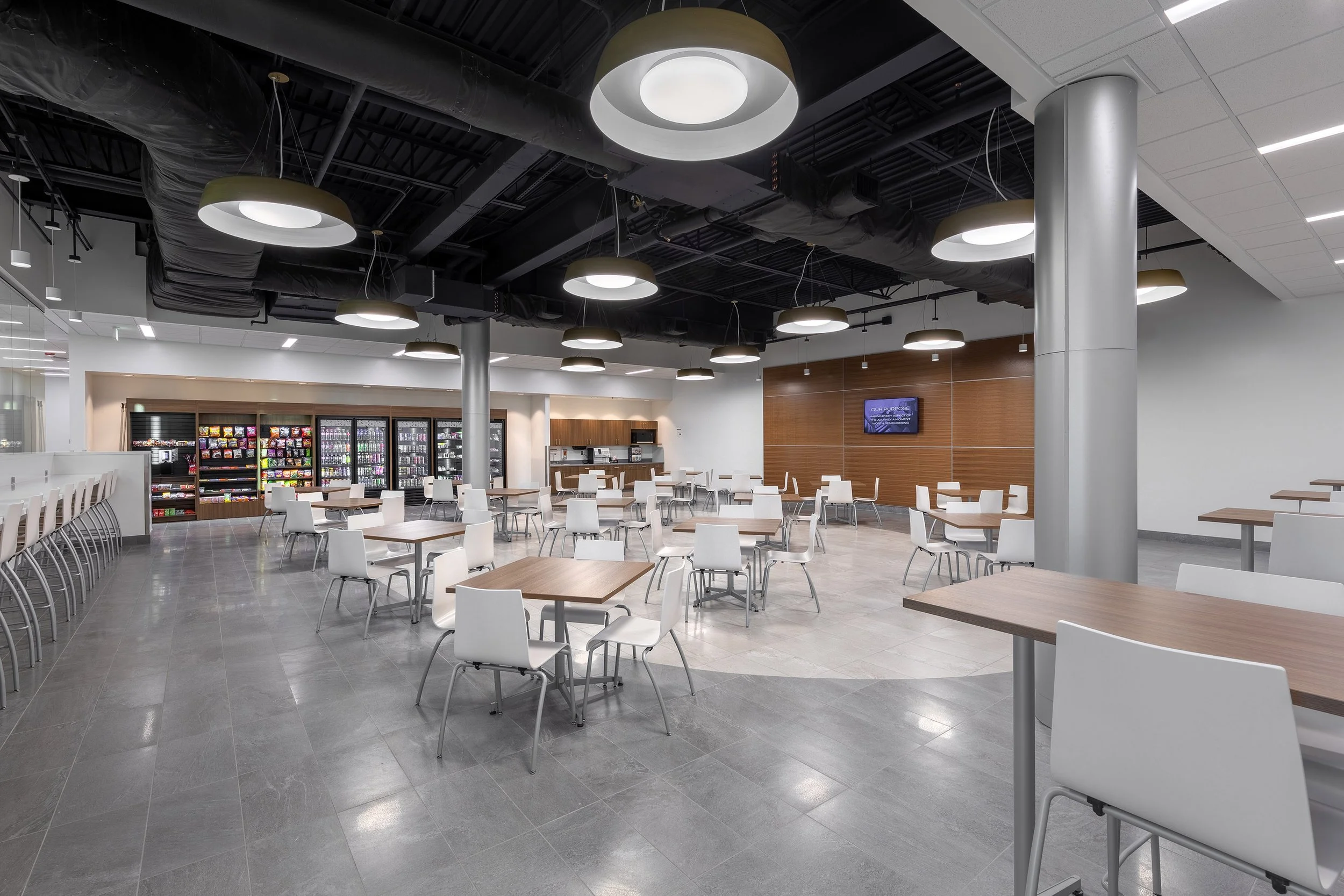 Modern cafeteria with white tables and chairs, vending machines along the back wall, and large circular ceiling lights. The clean, open space features gray flooring and a few people—its design inspired by Arizona’s innovative construction style.