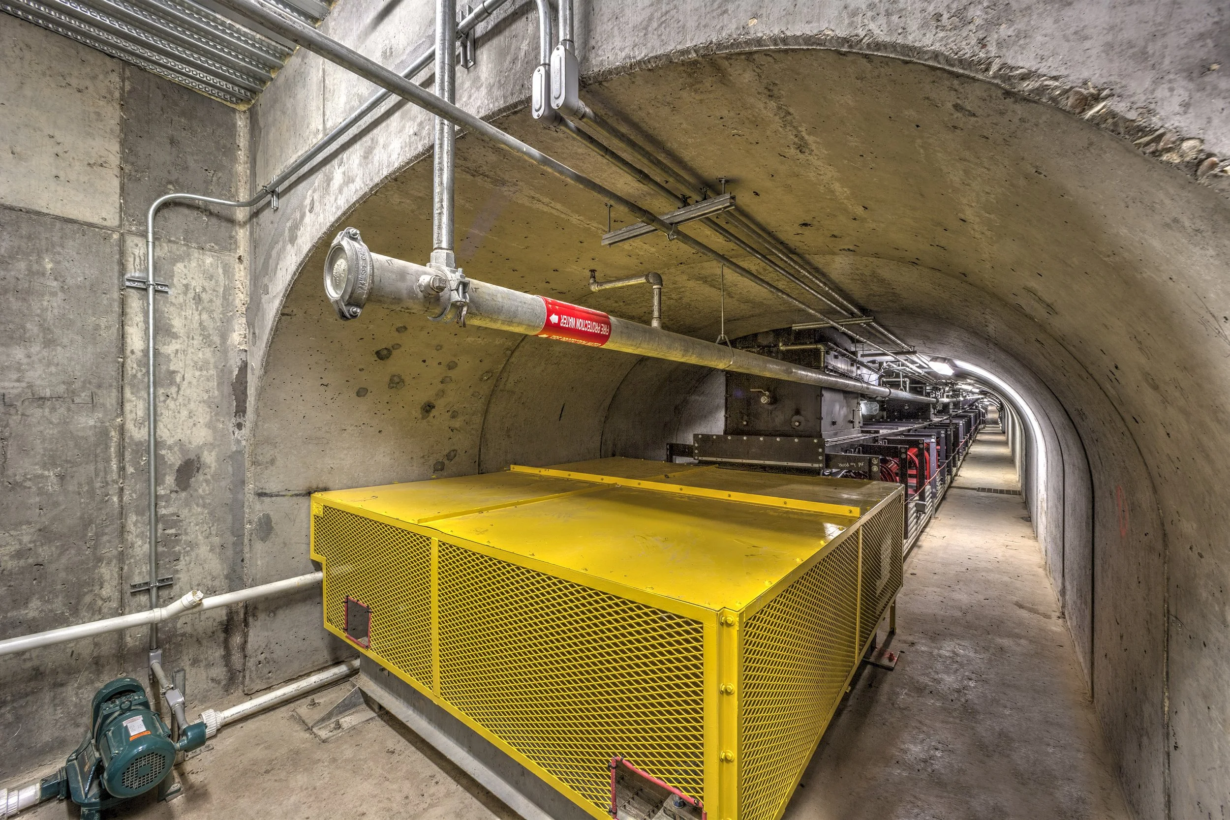 A concrete tunnel with yellow metal machinery, exposed pipes, and wires running along the walls and ceiling.