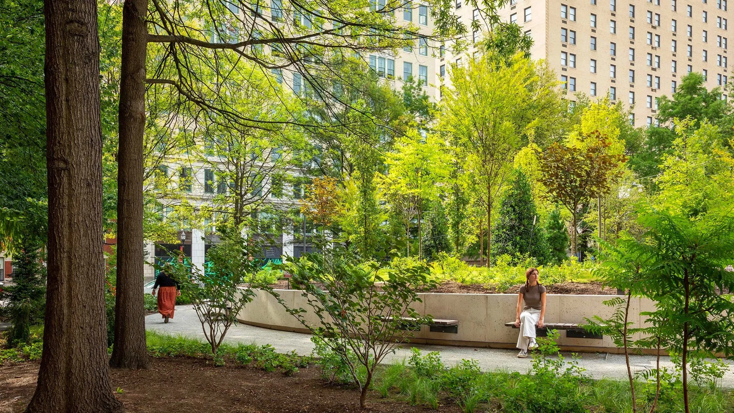 A woman sits on a concrete bench in a green park with trees and shrubs, with tall buildings in the background.