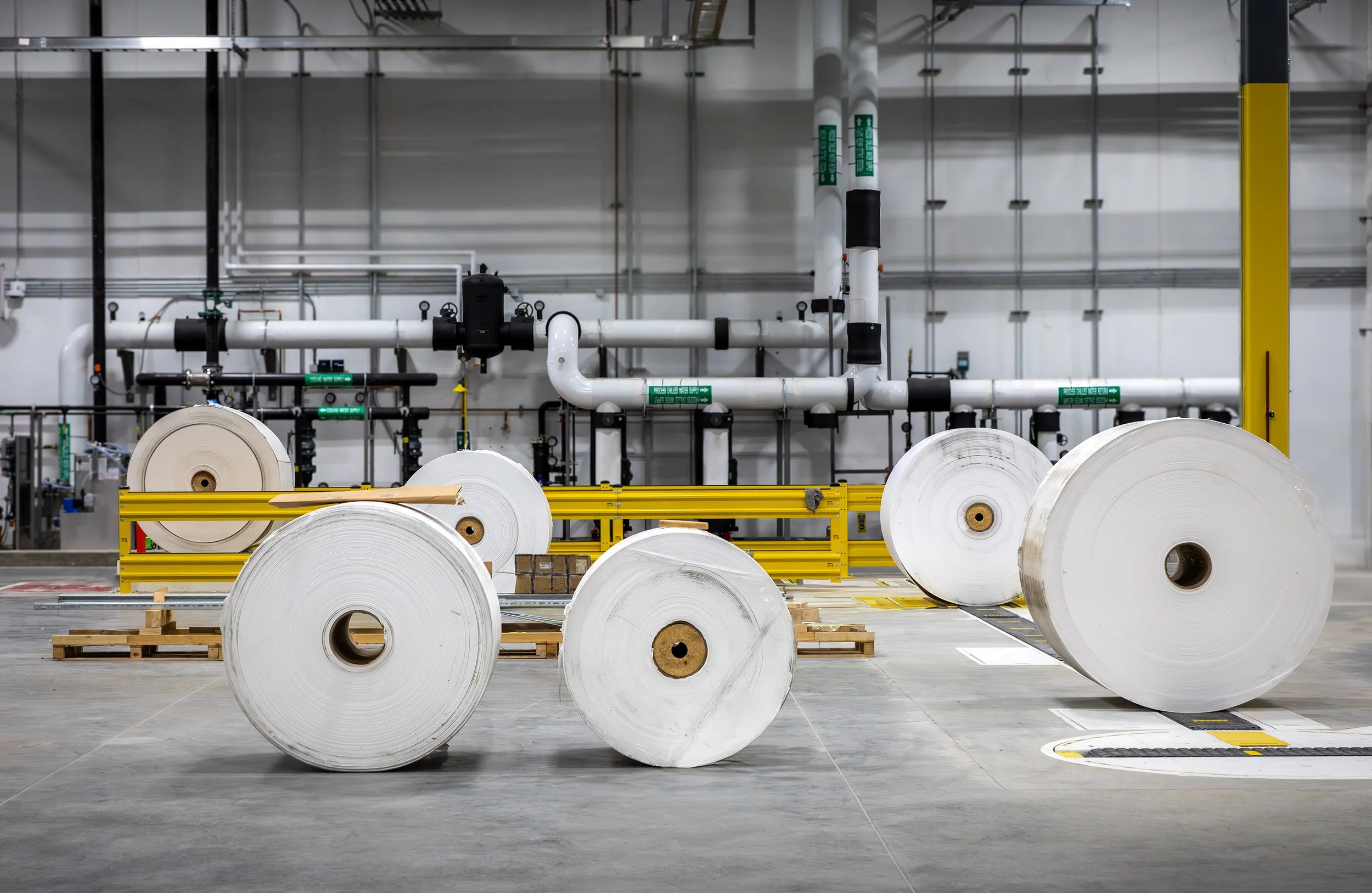 Large rolls of white industrial paper from Georgia Pacific are arranged on pallets inside a Dixie manufacturing facility with safety barriers and pipes.