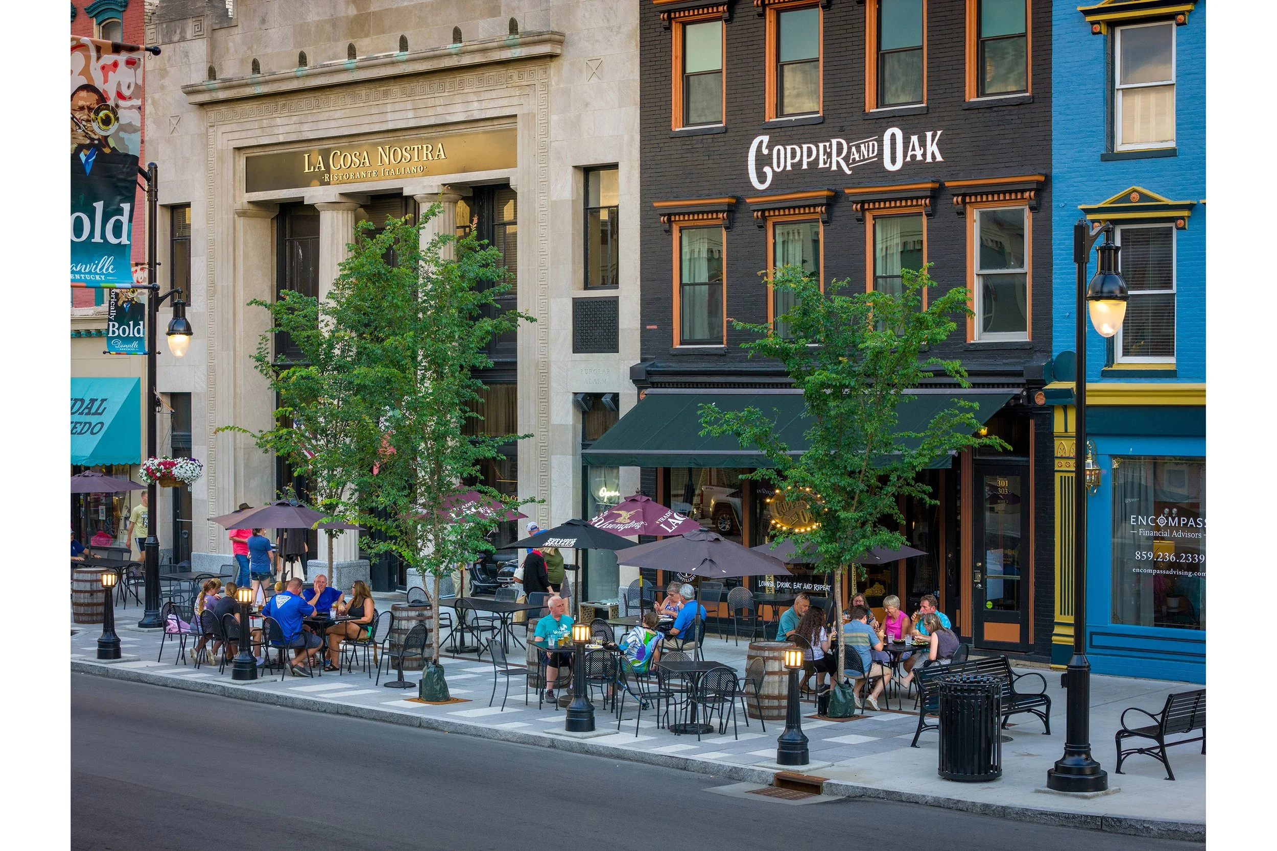 People are dining at outdoor tables along a vibrant streetscape, surrounded by historic shops and restaurants on a sunny day.