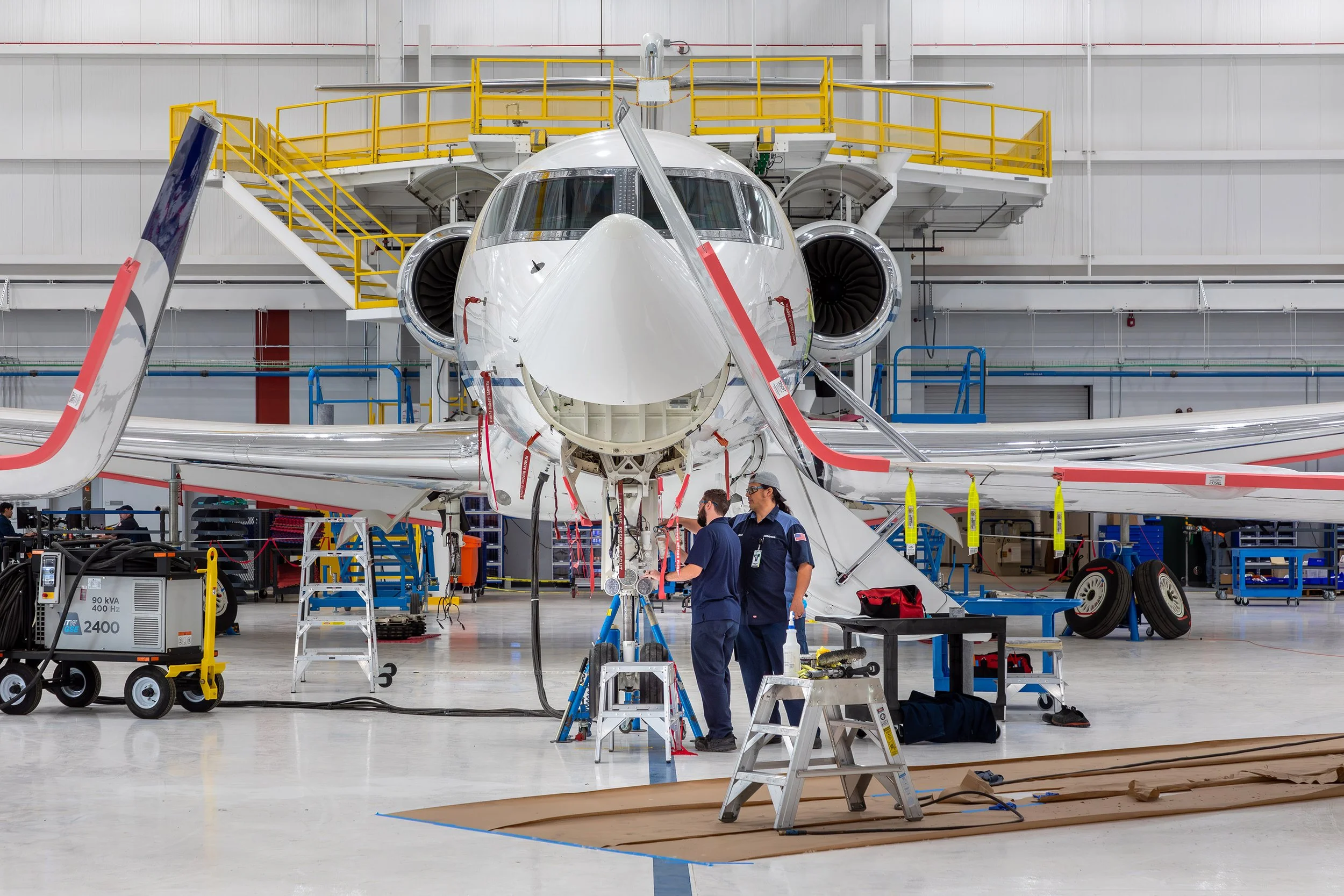 Two technicians work on the nose of a white jet inside a spacious aircraft hangar, showcasing the architectural details and organized maintenance equipment. The aircraft’s engines and wings are visible, with yellow railings on elevated walkways behin