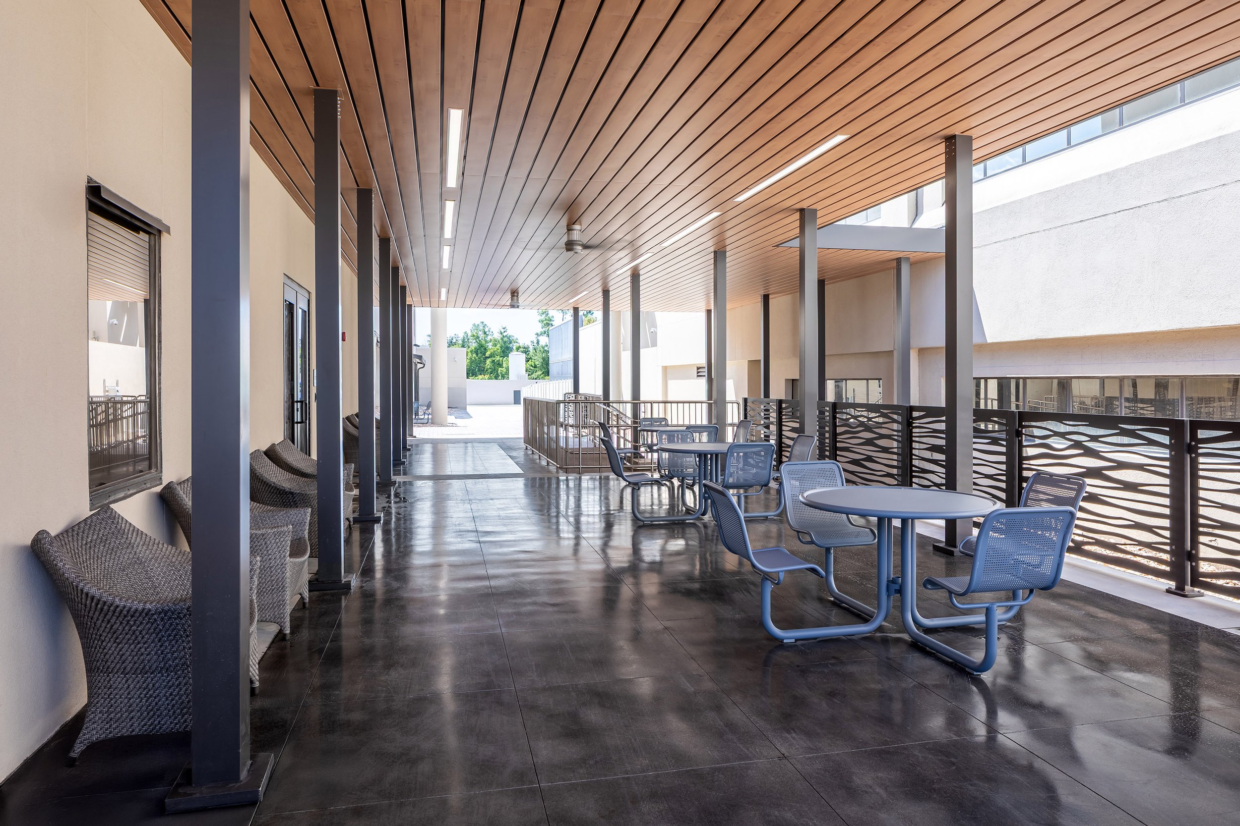 A modern building entrance with a wooden ceiling, metal columns, decorative railing, glass doors, and a wheelchair-accessible ramp creates an inviting outdoor setting under a clear blue sky—perfect for photography.